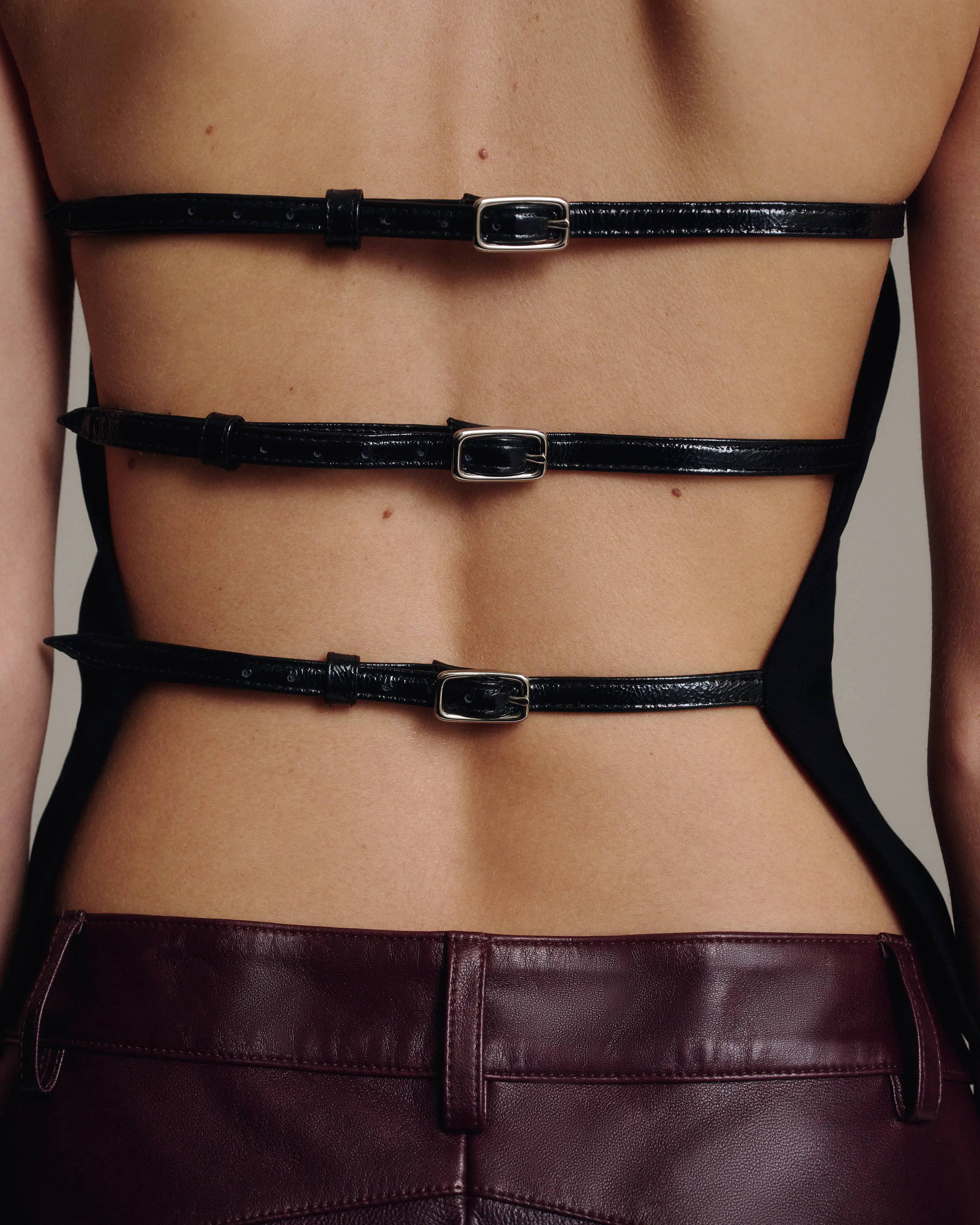 Close-up of the open back of a black halter top with three horizontal buckled leather straps, above the waistband of burgundy leather trousers, against a taupe background