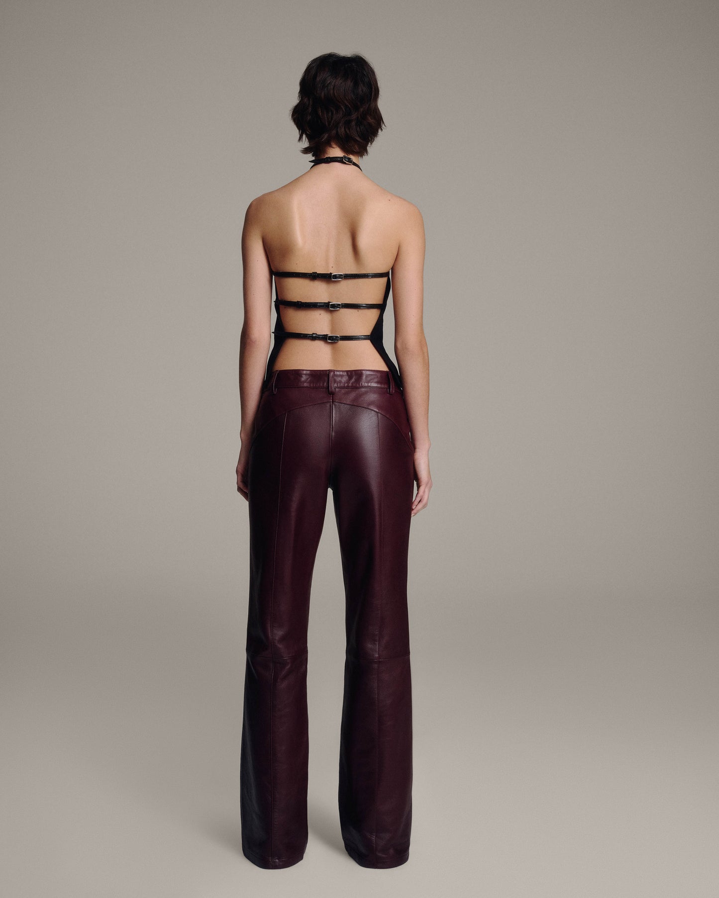 Back view of a model in a black halter top with three horizontal buckled straps across the open back, paired with burgundy leather trousers, full-length against a taupe background