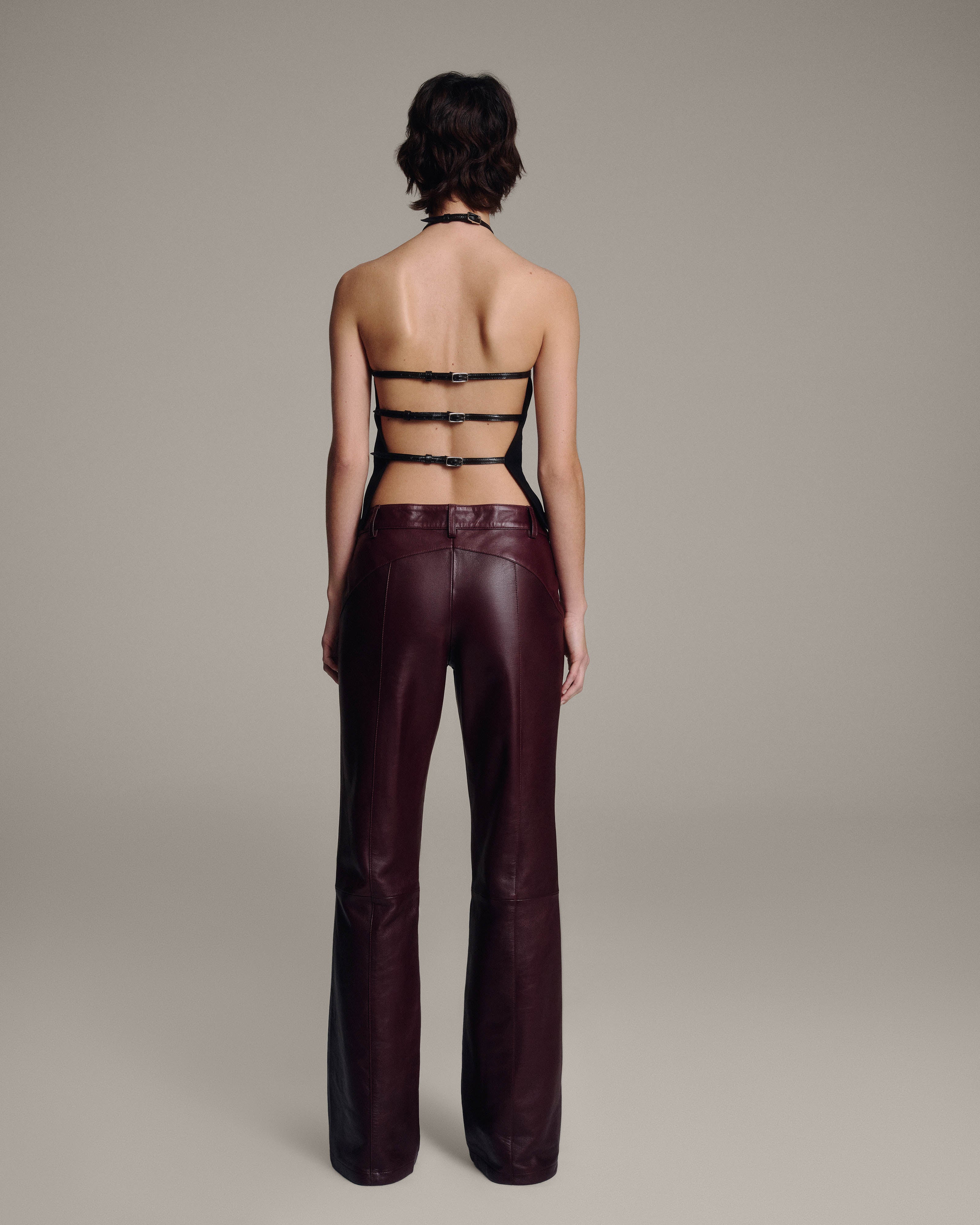 Back view of a model in a black halter top with three horizontal buckled straps across the open back, paired with burgundy leather trousers, full-length against a taupe background