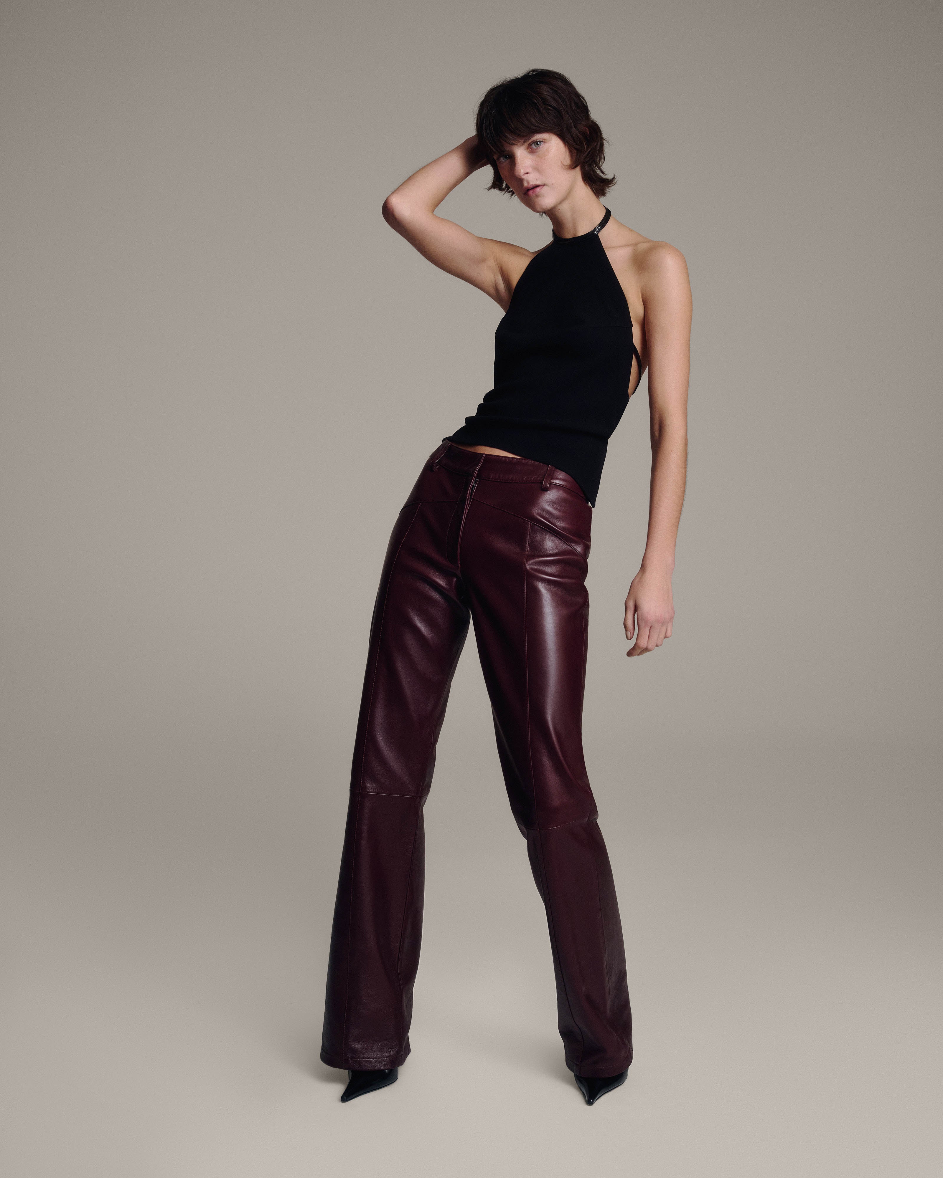 Model with one hand raised to her head wearing a black halter top and burgundy leather flared trousers, full-length angled view against a taupe background