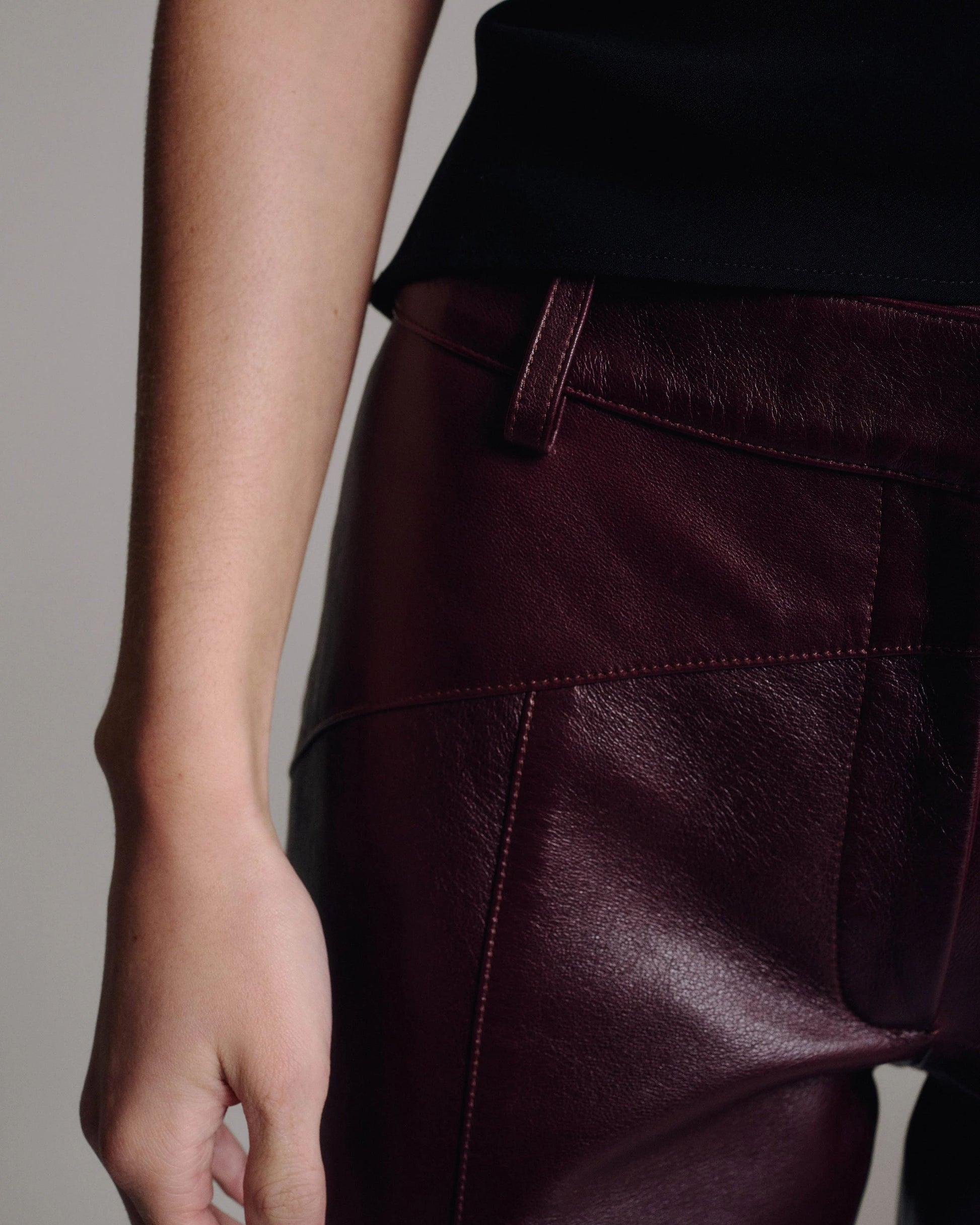 Close-up of the waistband, belt loop and pocket detail on burgundy leather trousers against a taupe background