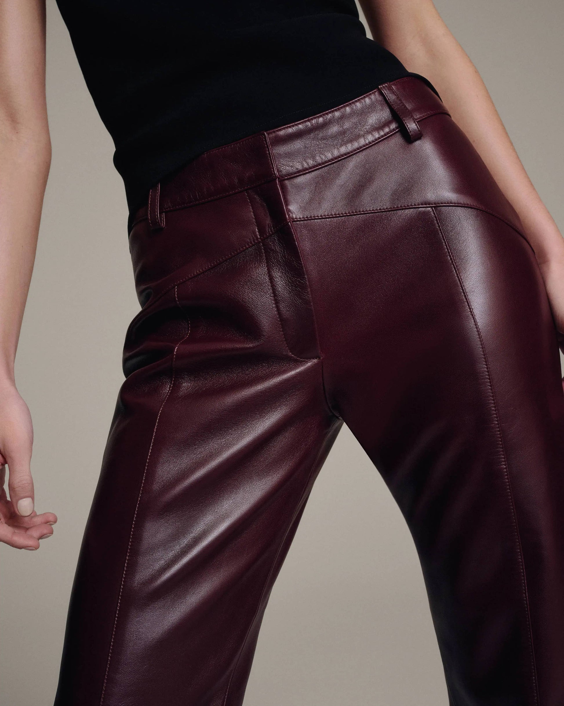 Model in low-rise burgundy leather trousers with a black top, cropped front view of the waist and hips against a taupe background