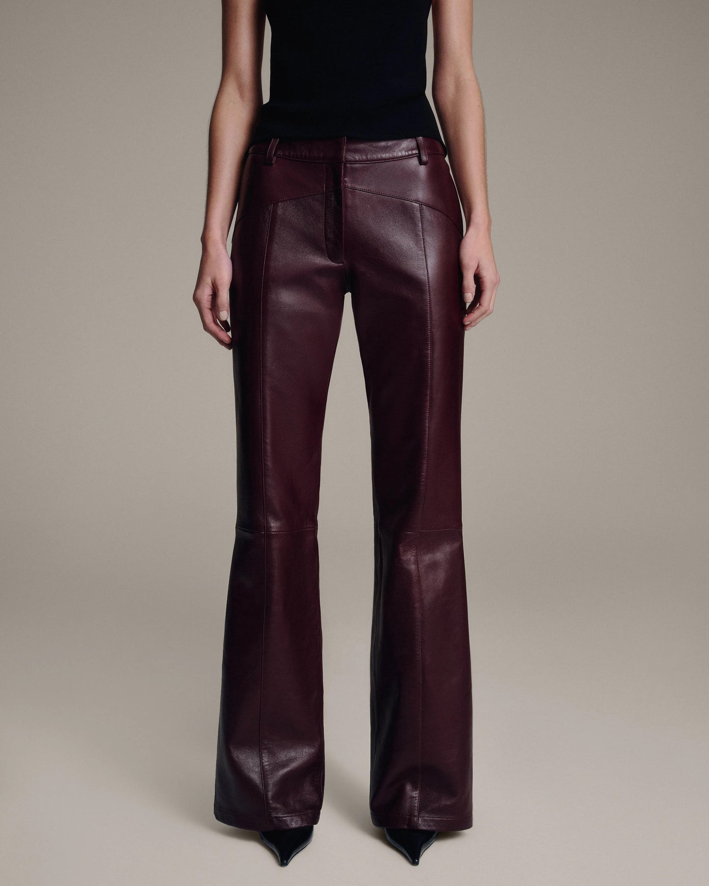 Model wearing burgundy leather flared trousers with topstitched seams, paired with a black top, lower-body front view against a taupe background
