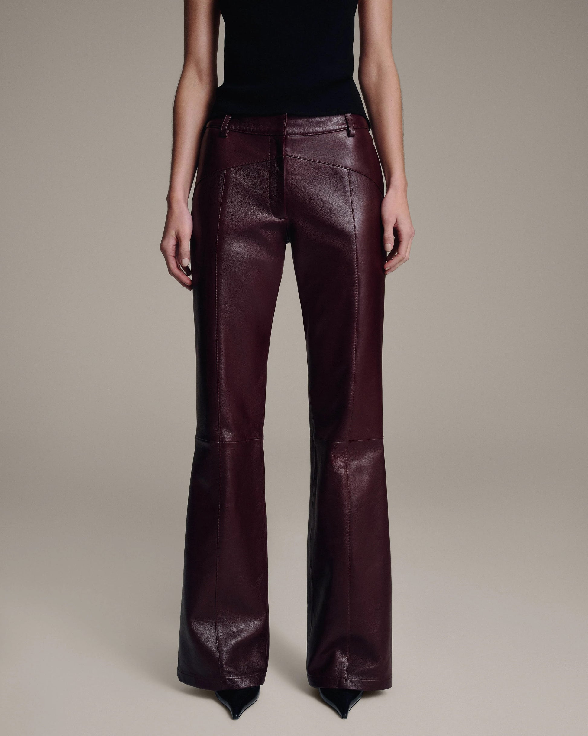 Model wearing burgundy leather flared trousers with topstitched seams, paired with a black top, lower-body front view against a taupe background