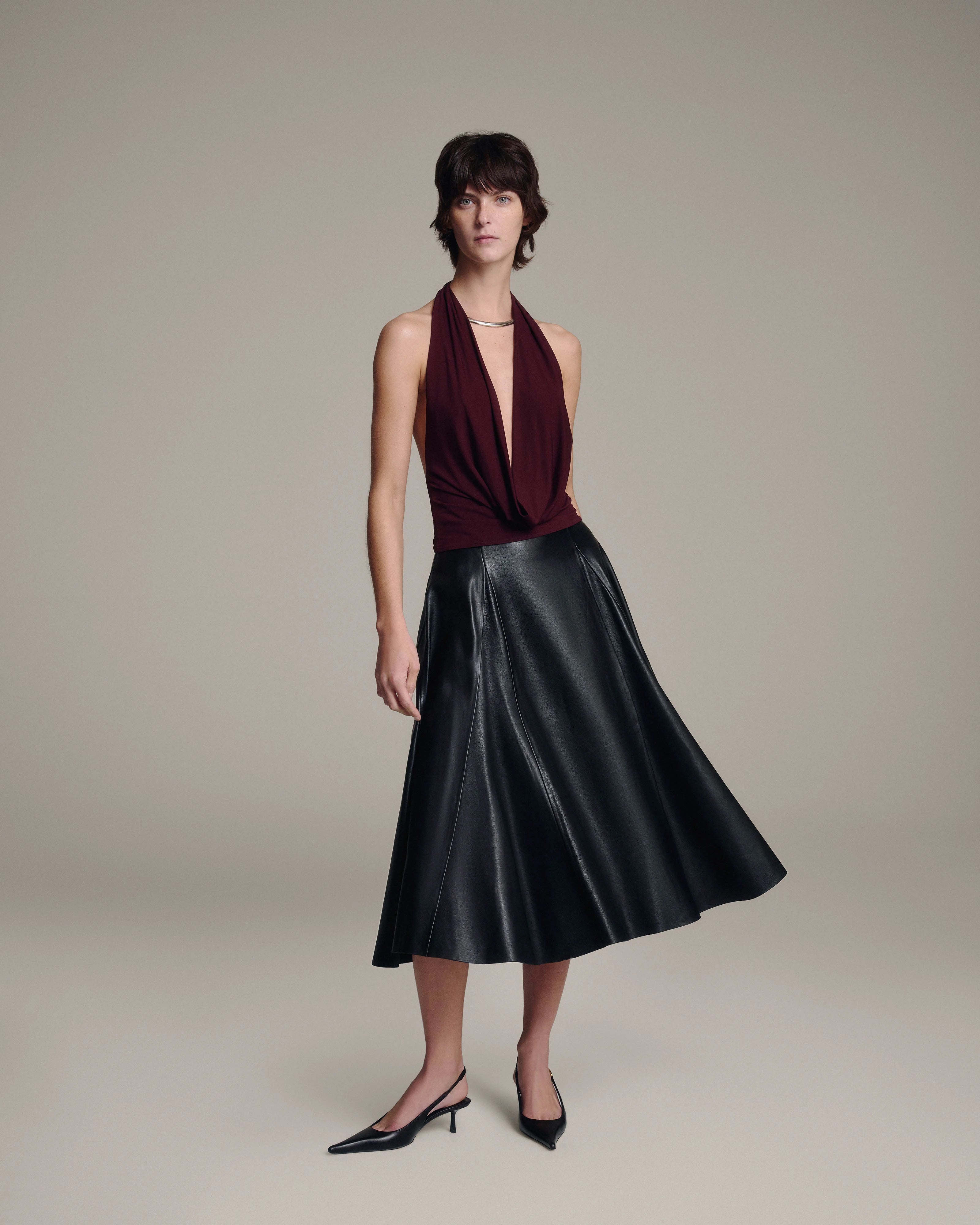A model wearing a sleeveless halter-neck dress with a deep V-neck in dark burgundy and a black leather skirt, paired with black pointed-toe heels, against a solid beige studio background.