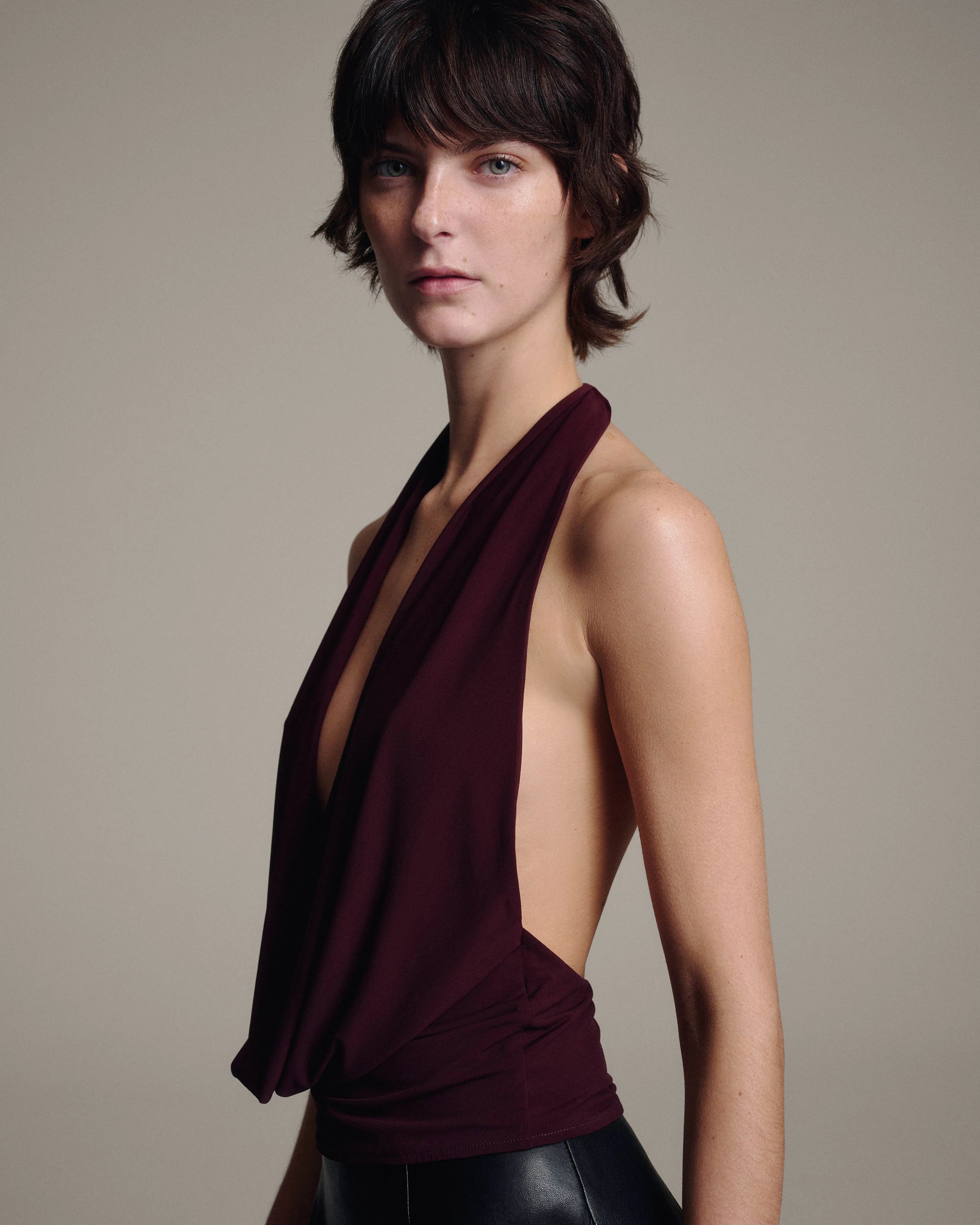 A model wearing a deep burgundy halter-neck wrap top with a dramatic side drape and open back, paired with a black leather skirt, set against a plain beige studio background.