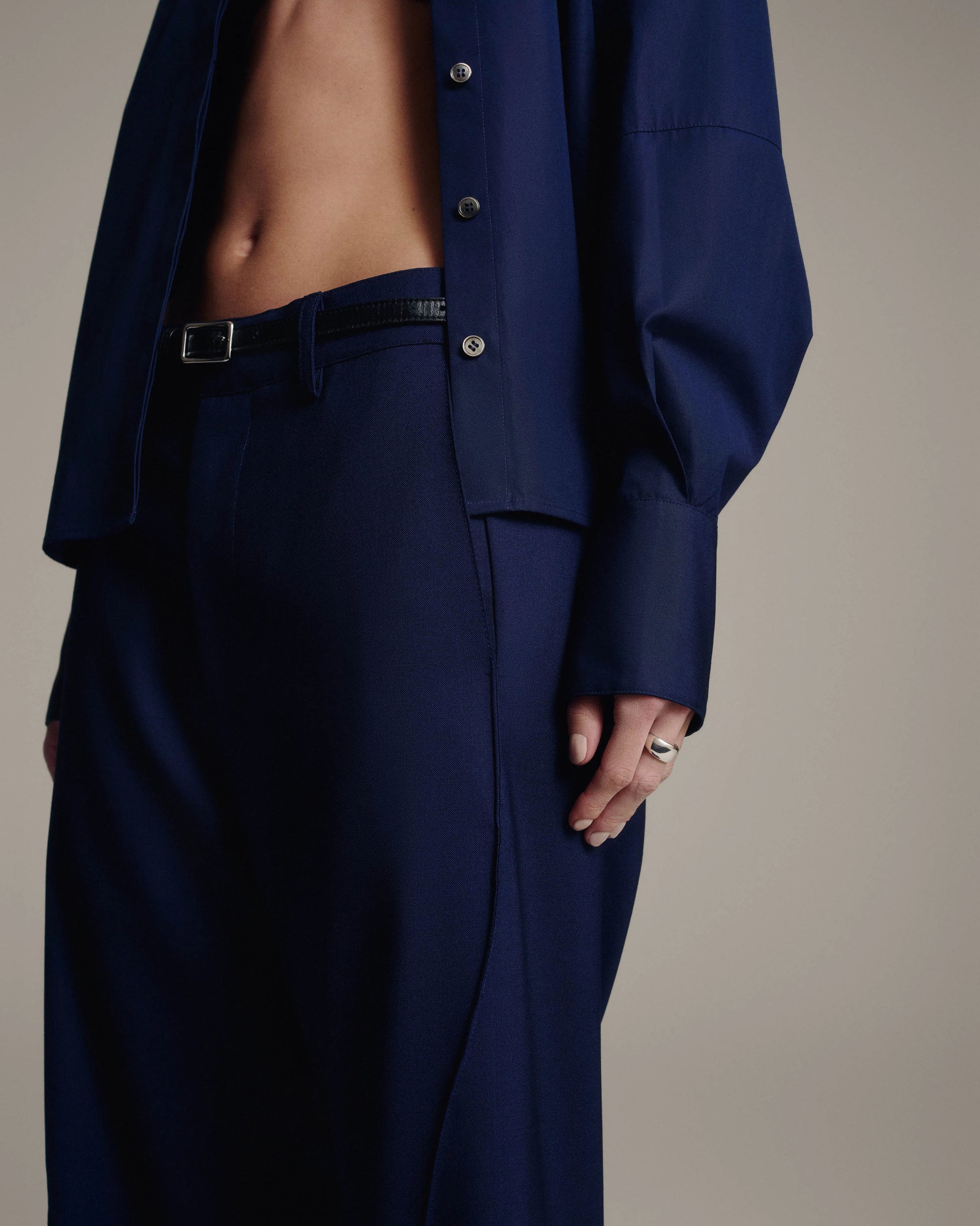 A close-up of a model wearing a navy blue tailored suit, featuring a cropped button-up jacket and matching trousers, with a black belt and silver buckle.