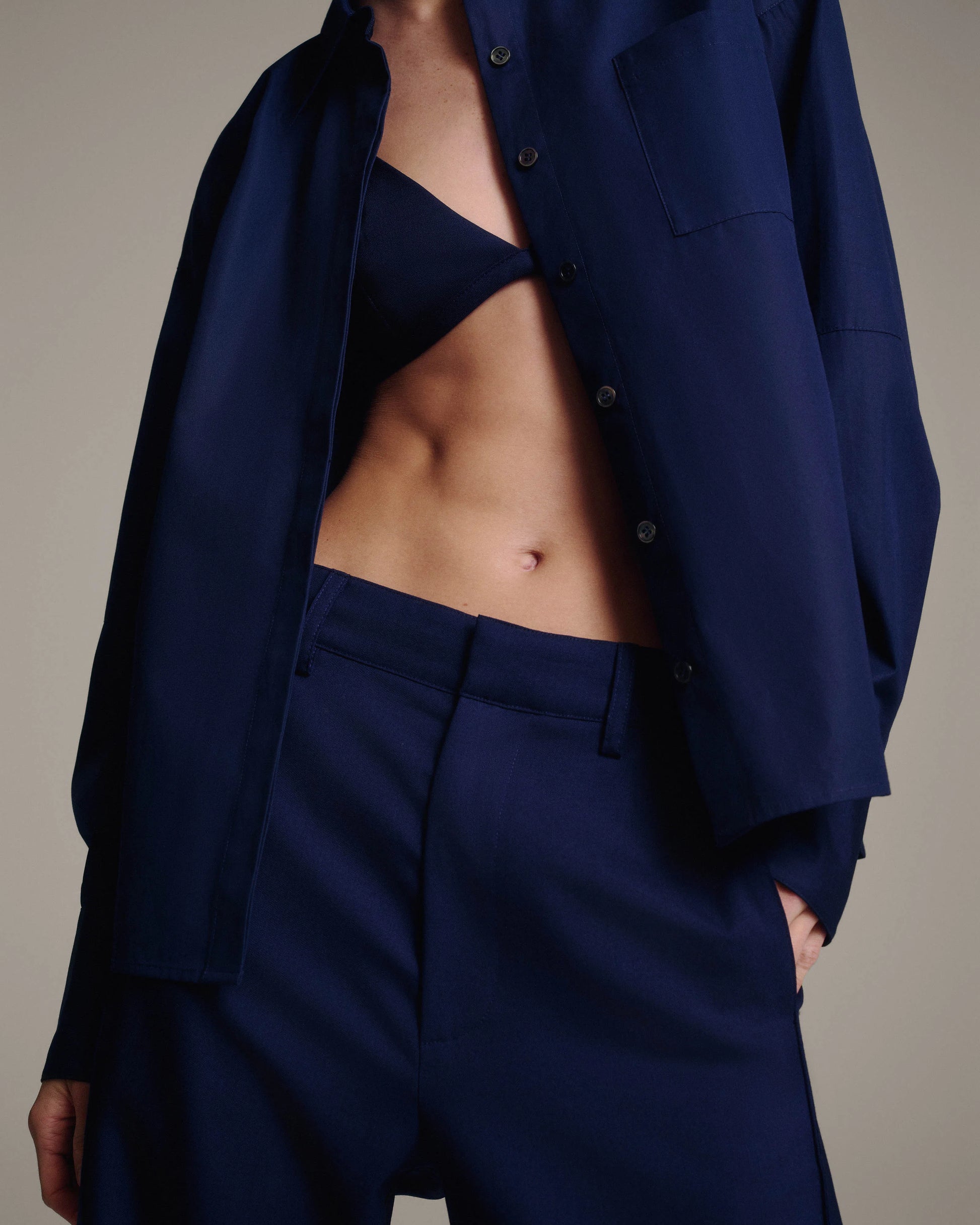 A close-up of a model wearing a matching navy blue set, featuring an oversized button-down shirt and high-waisted trousers, styled with a matching navy bra top.