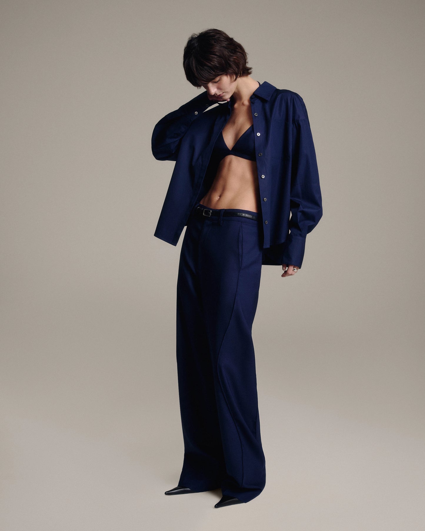 A model wearing a dark navy blue matching set consisting of a button-down shirt and wide-leg trousers, a navy bralette, and black pointed shoes against a neutral beige studio background.