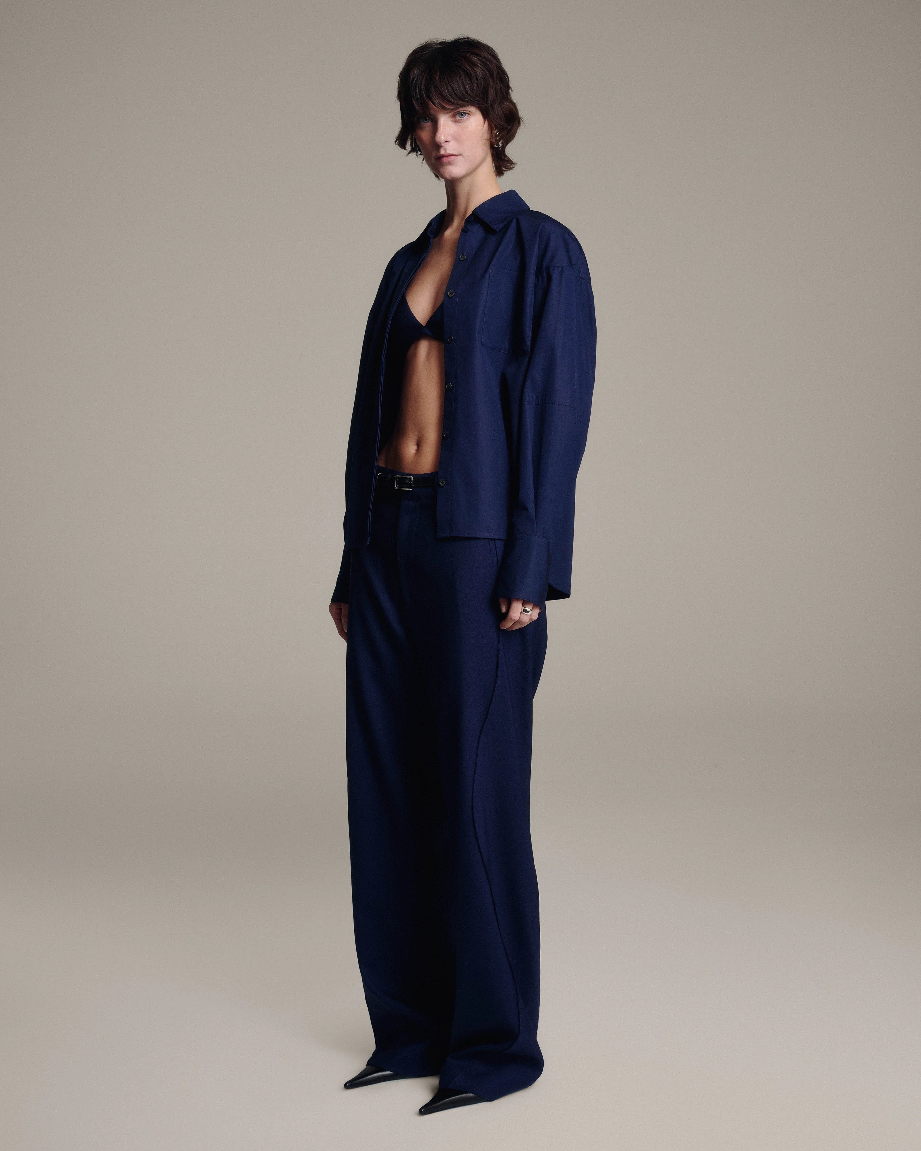 A model stands in a studio wearing a dark navy blue two-piece suit consisting of an open long-sleeve blazer, matching wide-leg trousers, bralette, and pointed black shoes against a neutral beige background.