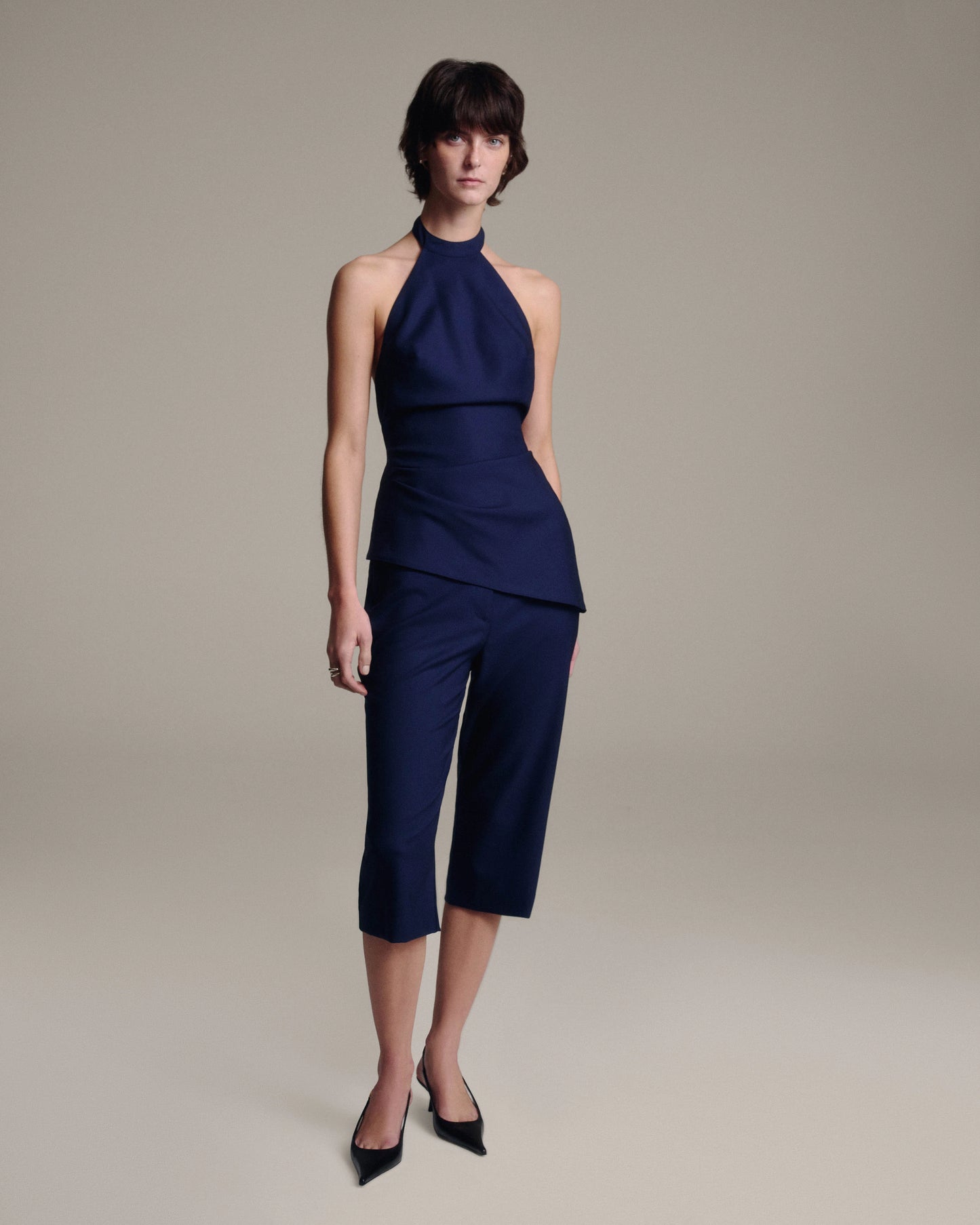 A model wearing a navy blue sleeveless halter top and matching cropped capri pants, paired with black pointed-toe heels, against a solid warm gray studio background.