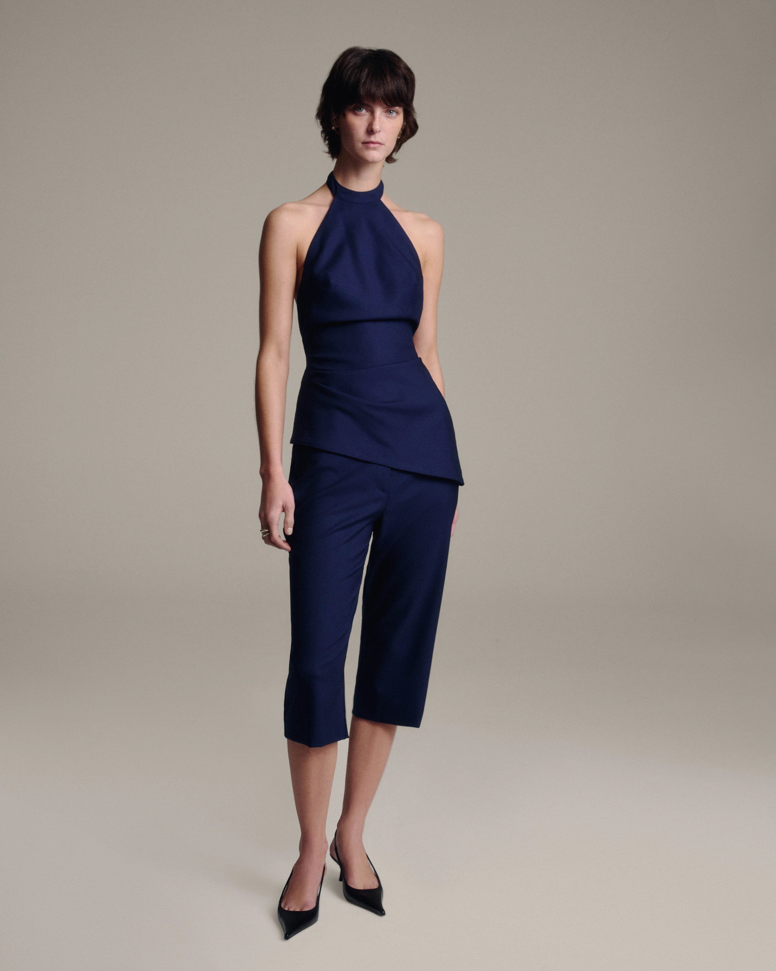 A model wearing a navy blue sleeveless halter top and matching cropped capri pants, paired with black pointed-toe heels, against a solid warm gray studio background.