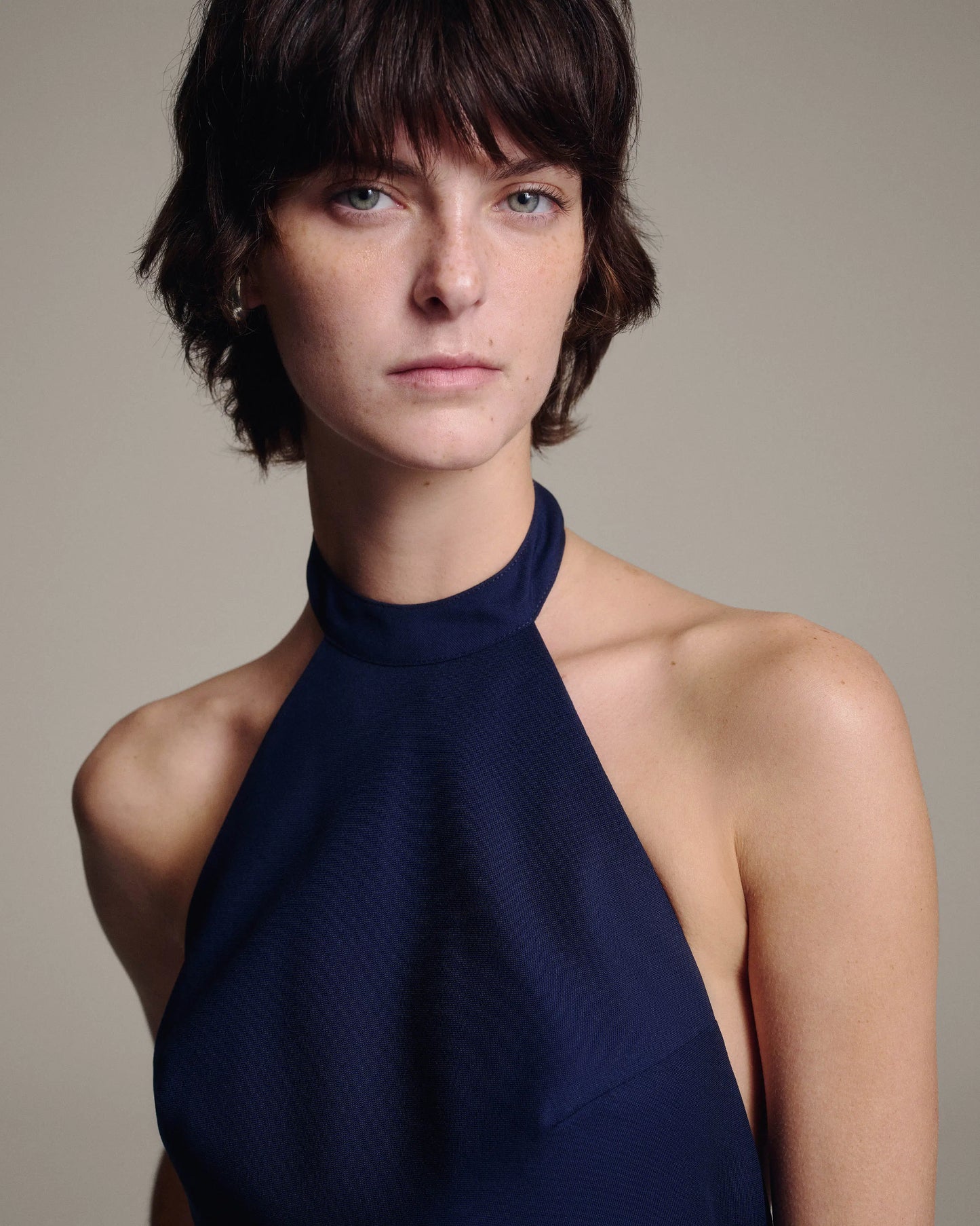 A close-up portrait of a model with short dark hair wearing a dark navy blue halter-neck top against a neutral beige background.