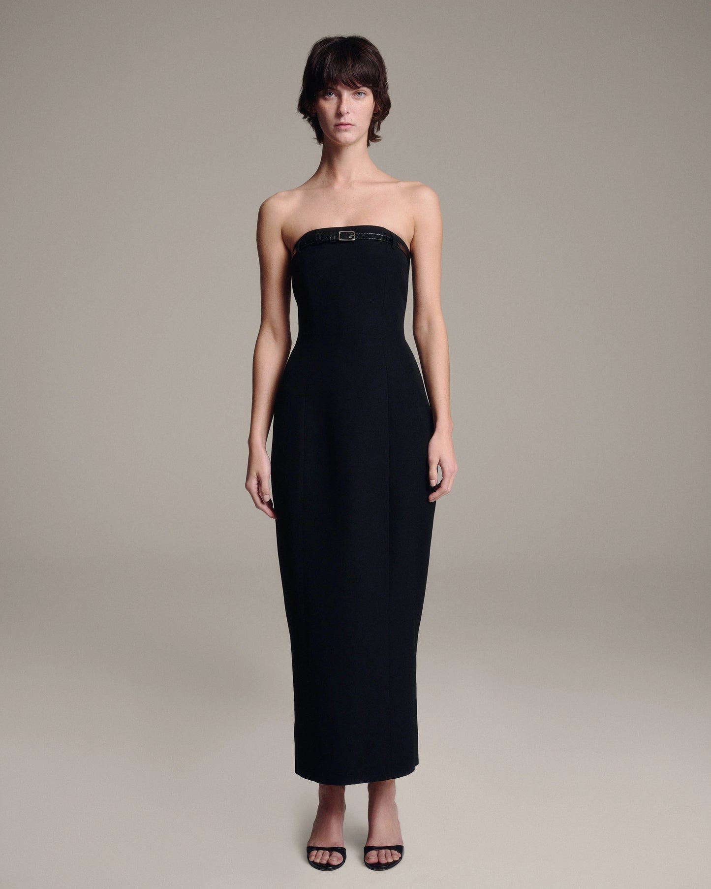 A woman modeling a strapless black midi dress with a structured bodice and a small belt at the bust, photographed against a plain warm gray studio background.