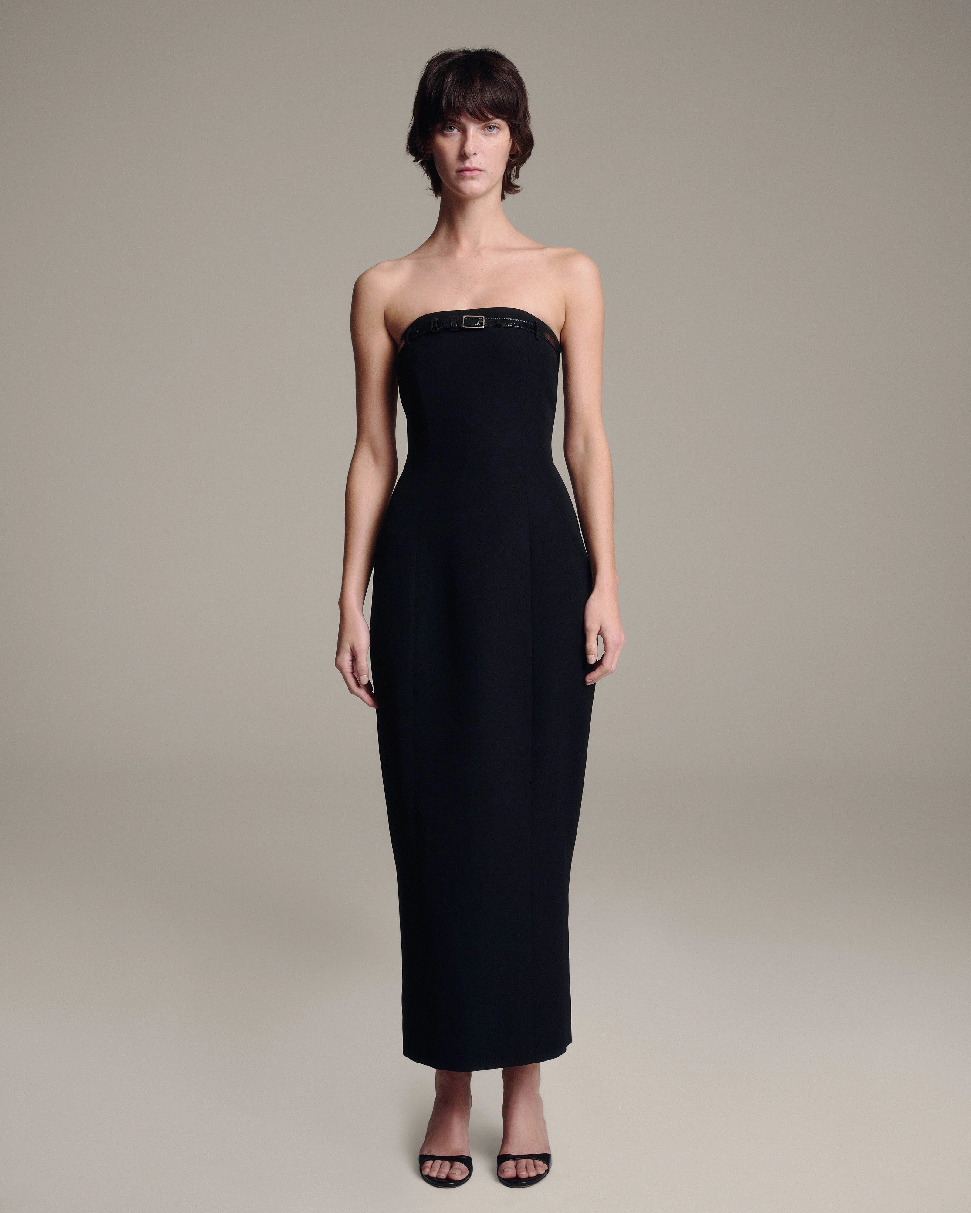 A woman modeling a strapless black midi dress with a structured bodice and a small belt at the bust, photographed against a plain warm gray studio background.