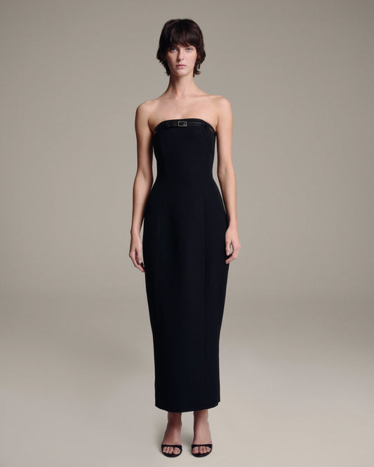 A woman modeling a strapless black midi dress with a structured bodice and a small belt at the bust, photographed against a plain warm gray studio background.