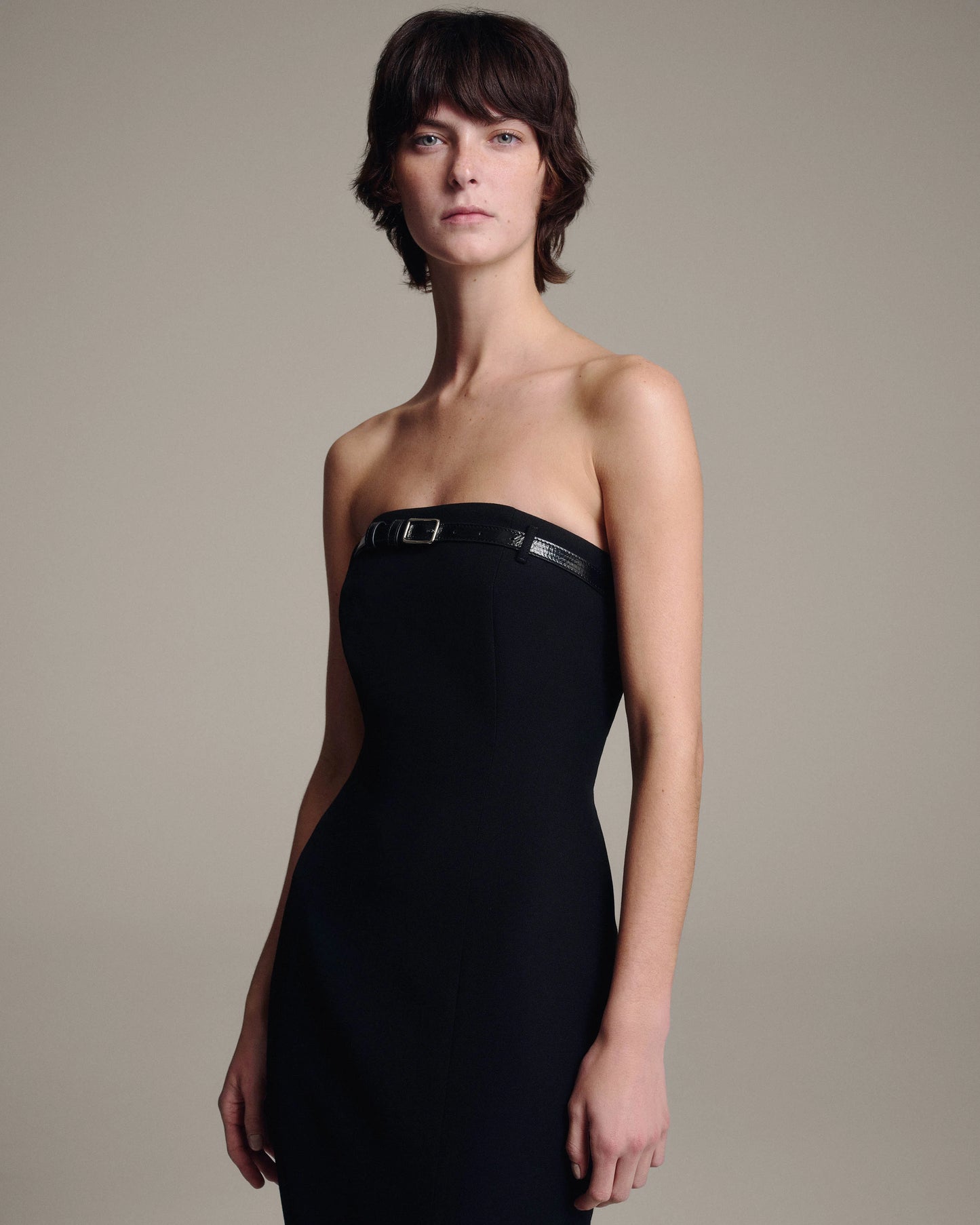A model wearing a black strapless dress with a thin black belt detail at the bust and a small buckle, photographed against a neutral beige studio background.