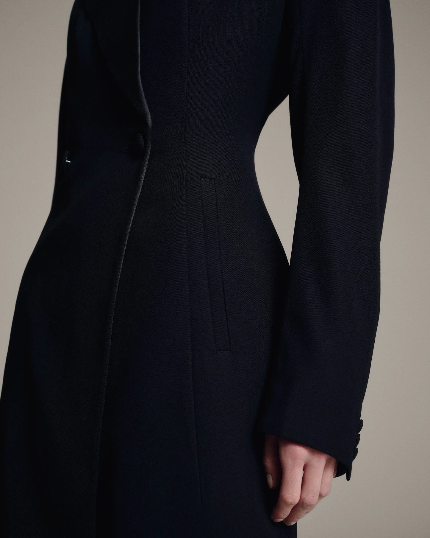 A close-up of a model wearing a long, structured black coat with a single-breasted button closure, a vertical welt pocket, and buttoned cuffs.