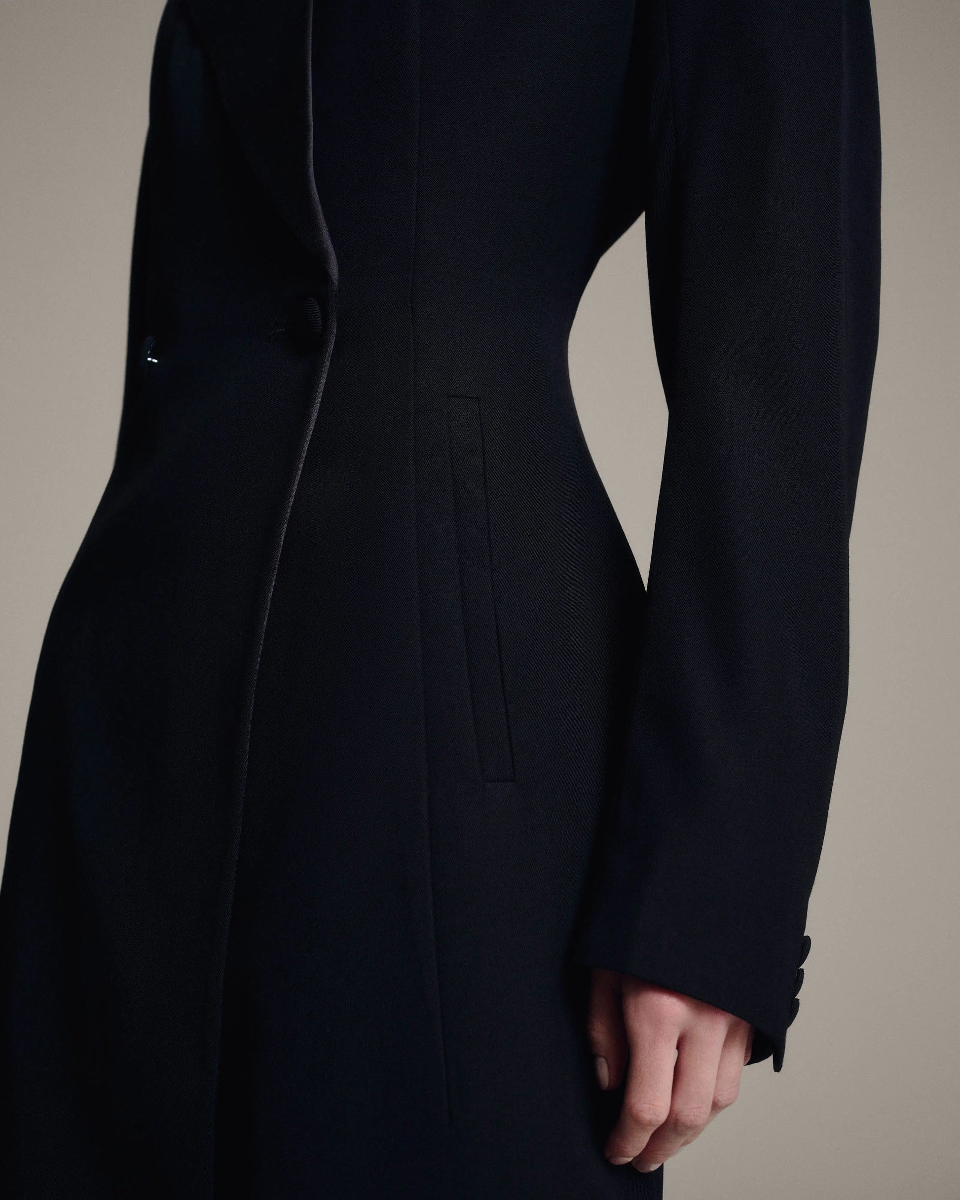 A close-up of a model wearing a long, structured black coat with a single-breasted button closure, a vertical welt pocket, and buttoned cuffs.