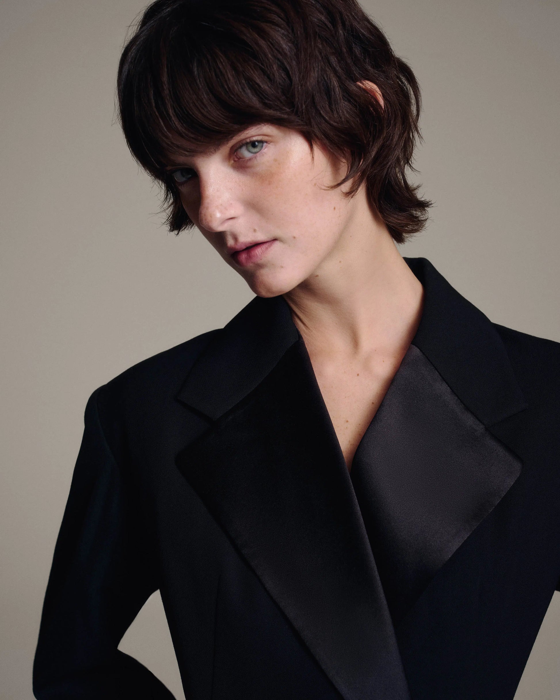 A close-up portrait of a model with short dark brown hair wearing a black double-breasted blazer with satin lapels, set against a solid warm gray background.