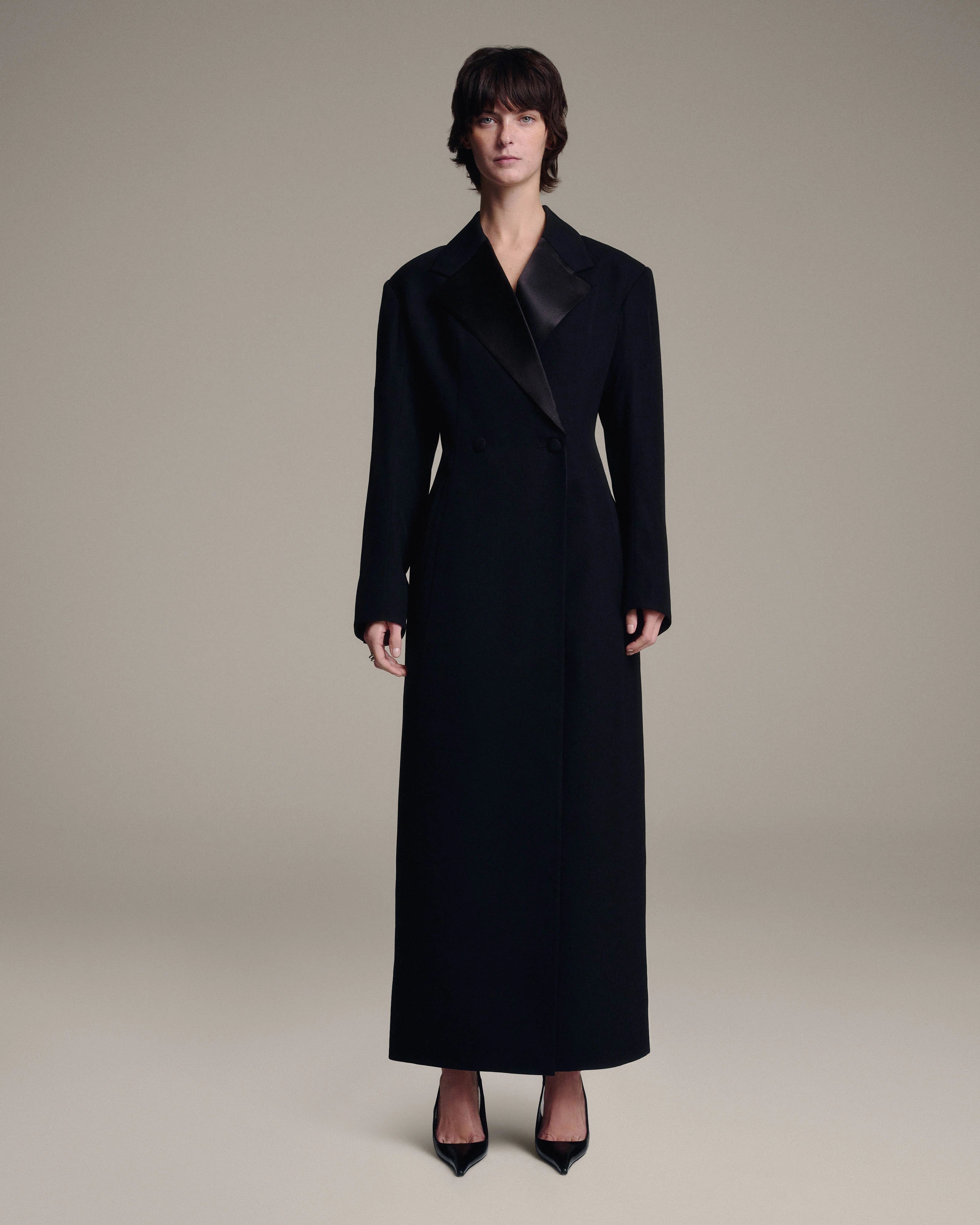 A model stands against a plain beige studio background wearing a long black coat with black satin lapels and black pointed-toe heels.