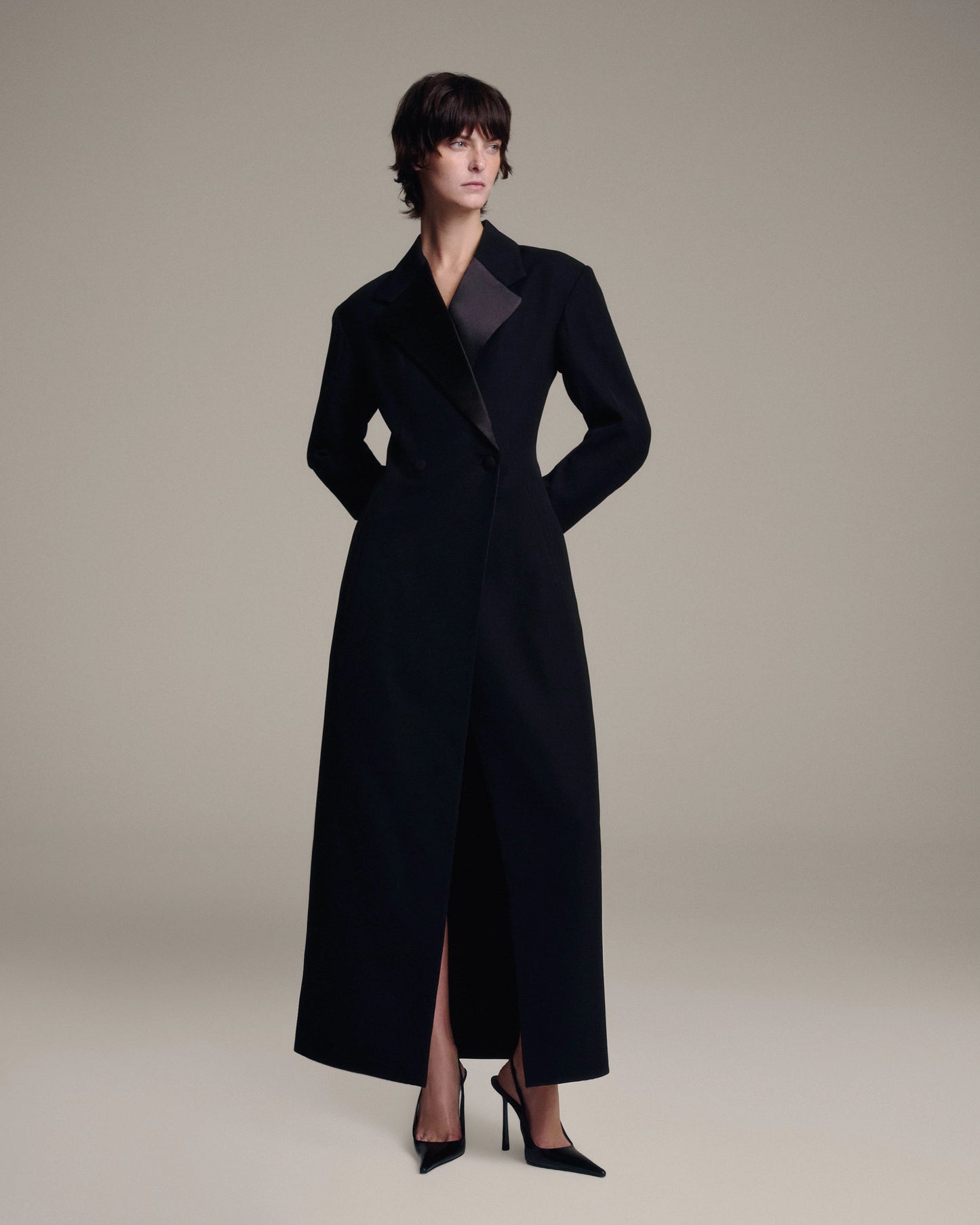A model wearing a long black tailored coat with satin lapels, paired with black high heels, photographed against a solid light beige studio background.