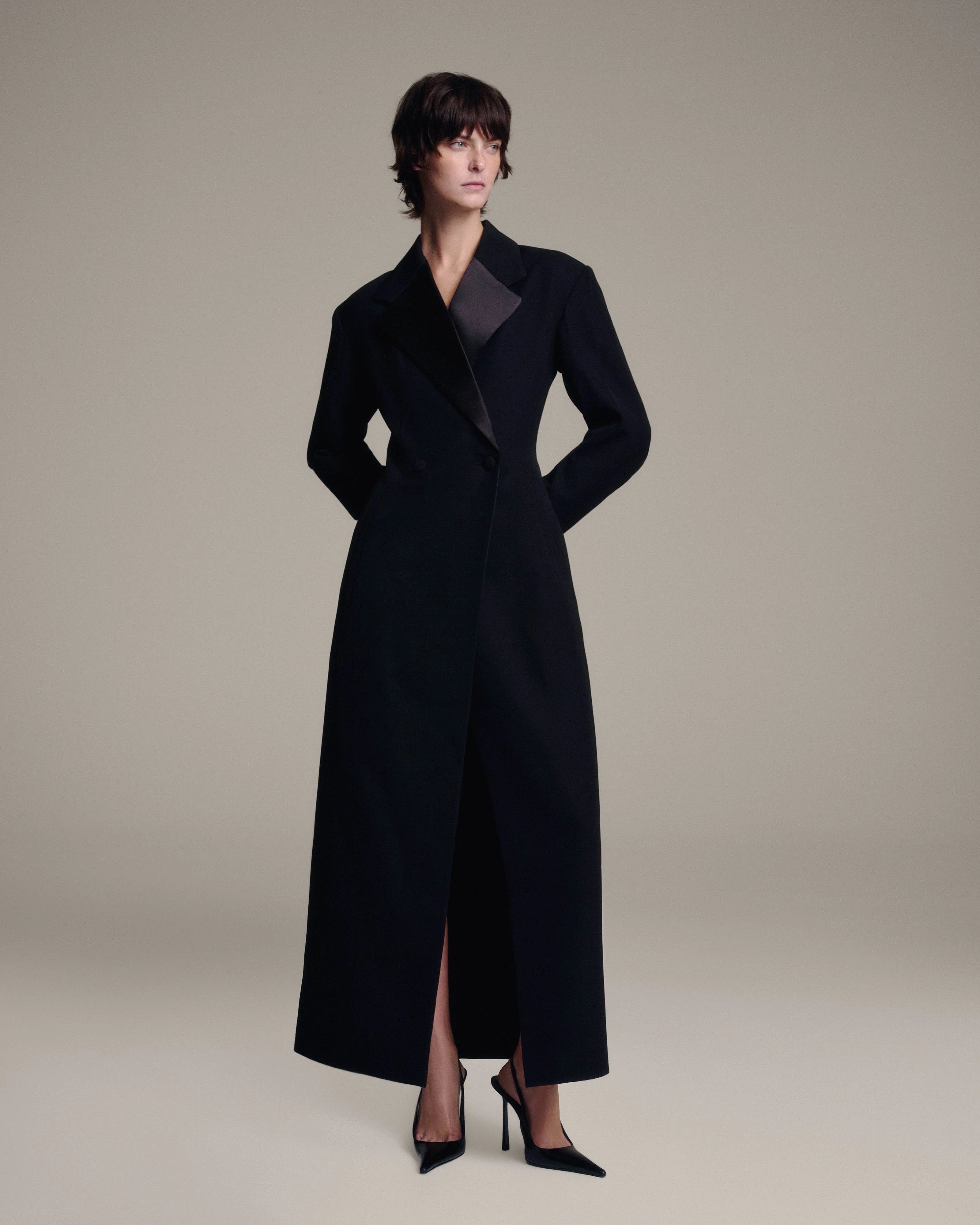 A model wearing a long black tailored coat with satin lapels, paired with black high heels, photographed against a solid light beige studio background.