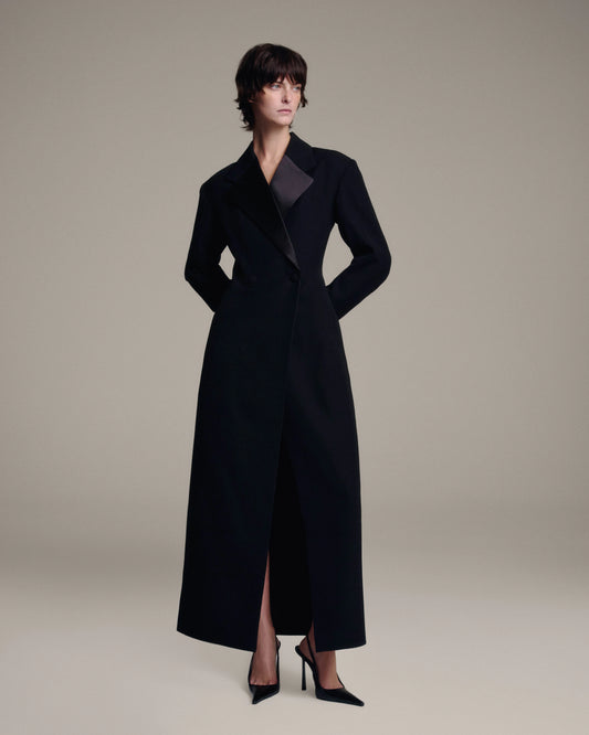 A model wearing a long black tailored coat with satin lapels, paired with black high heels, photographed against a solid light beige studio background.