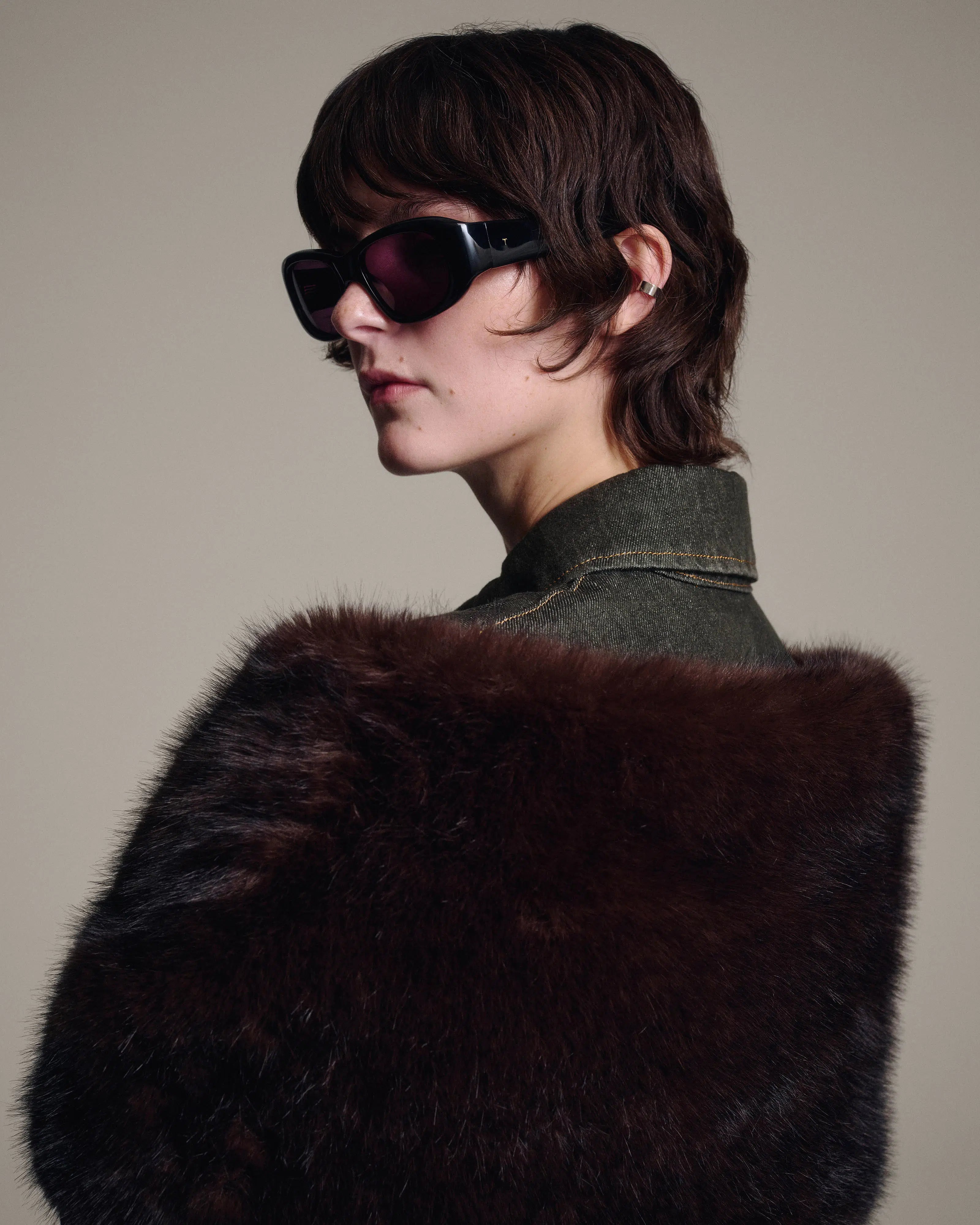 A model with short dark hair wearing black sunglasses and a dark brown fur stole over a dark denim jacket, photographed against a solid beige background.