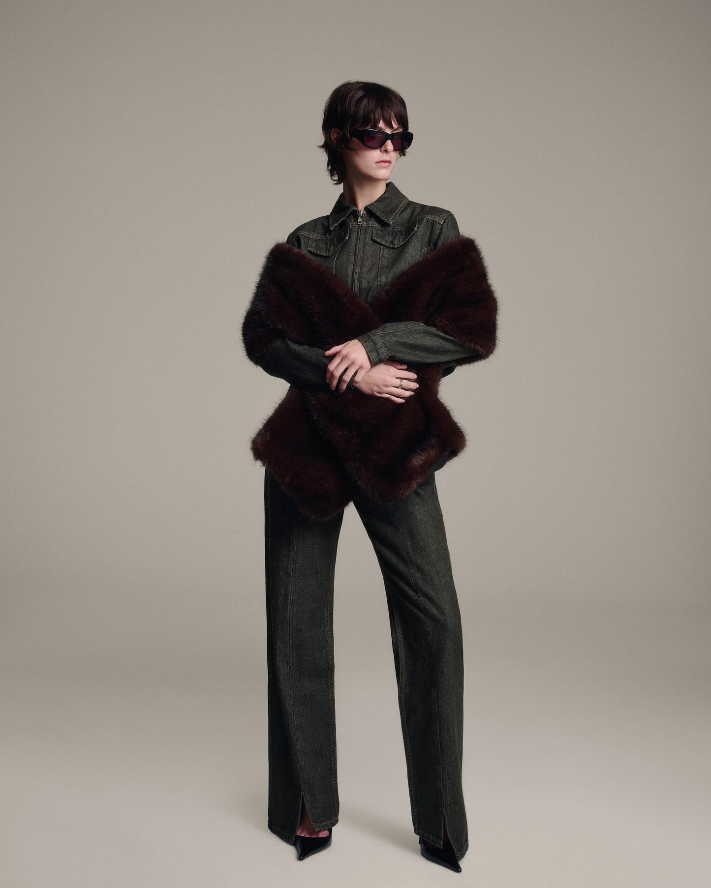 A model stands against a neutral beige studio background wearing a tailored denim jacket, matching wide-leg denim trousers, black pointed shoes, and a dark brown faux fur shawl. The model is also wearing dark sunglasses.
