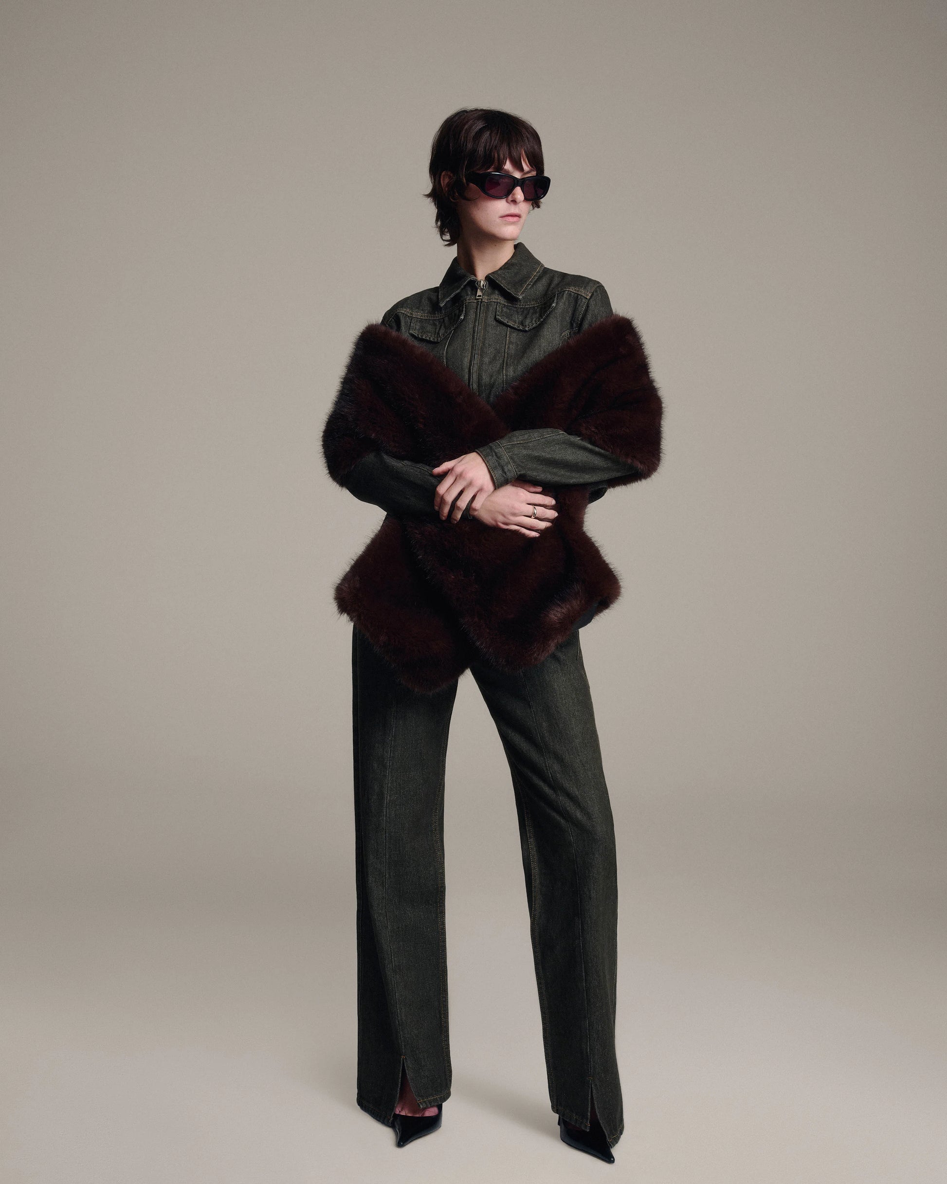 A model stands against a neutral beige studio background wearing a tailored denim jacket, matching wide-leg denim trousers, black pointed shoes, and a dark brown faux fur shawl. The model is also wearing dark sunglasses.