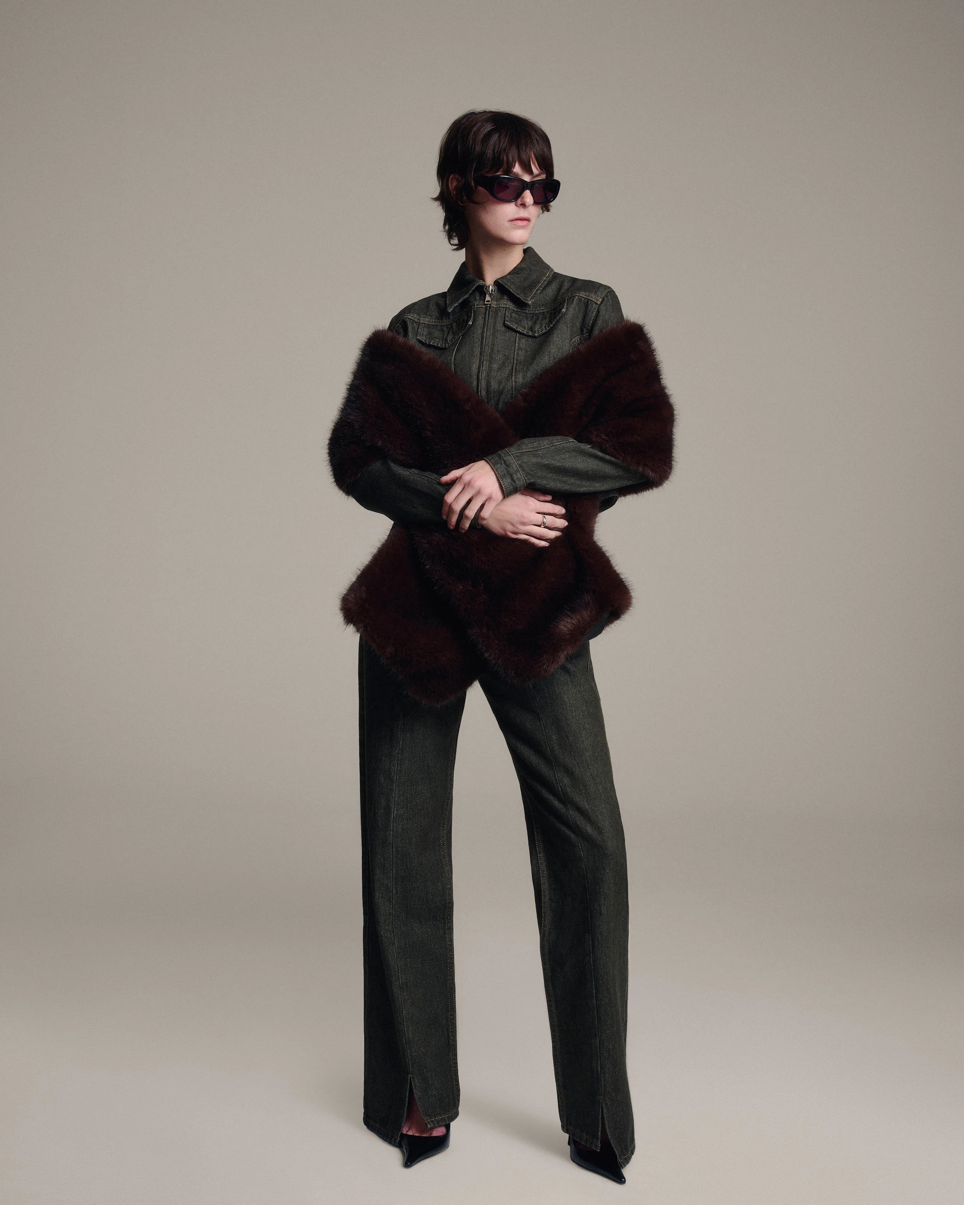 A model stands against a neutral beige studio background wearing a tailored denim jacket, matching wide-leg denim trousers, black pointed shoes, and a dark brown faux fur shawl. The model is also wearing dark sunglasses.