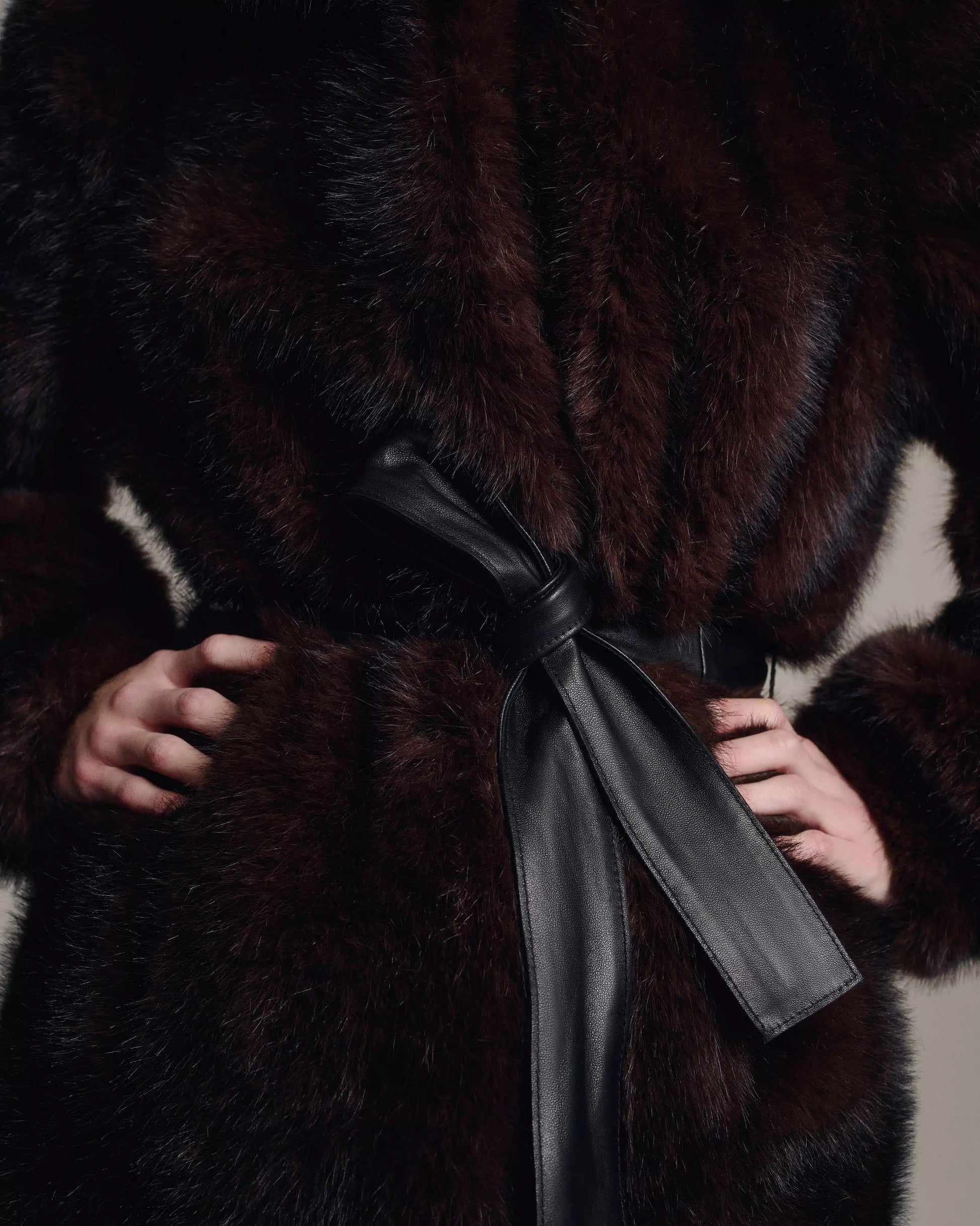 Close-up of a dark brown fur coat with a black leather belt tied at the waist, showing textured fur and a person's hands on their hips.