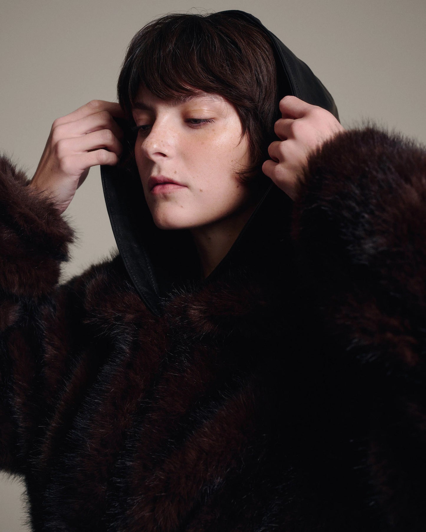 A model wearing a dark brown fur hooded coat, pulling the black leather hood over their head with both hands.