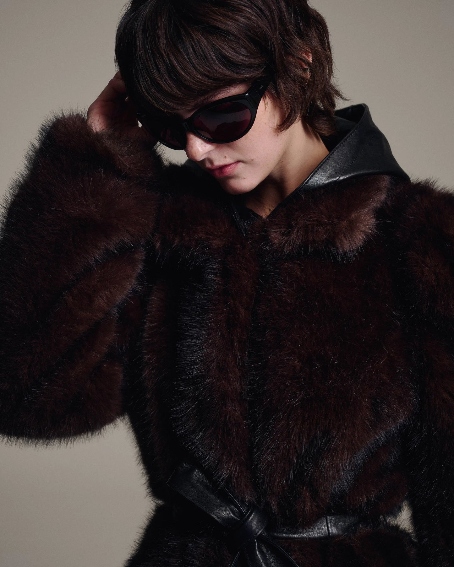 A model wearing a luxurious dark brown fur coat with black leather trim and a matching black leather belt, styled with dark sunglasses against a neutral beige background.