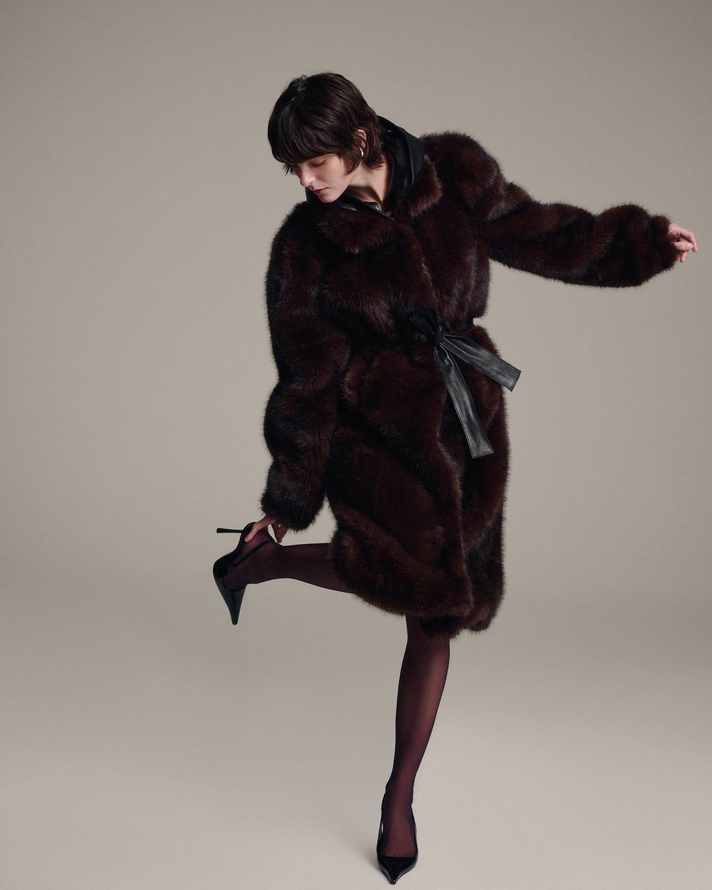 A model wearing a dark brown fur coat with a black leather belt, dark brown tights, and black pointed-toe heels, posing dynamically against a solid light gray studio background.
