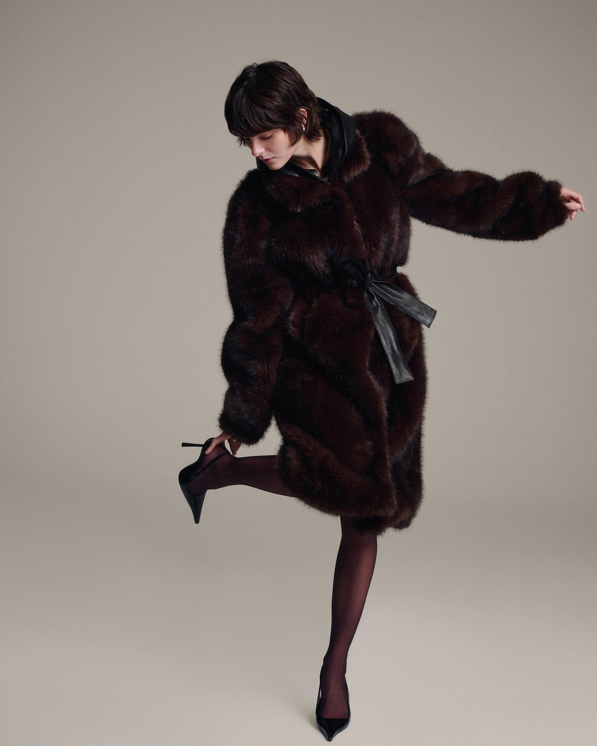 A model wearing a dark brown fur coat with a black leather belt, dark brown tights, and black pointed-toe heels, posing dynamically against a solid light gray studio background.