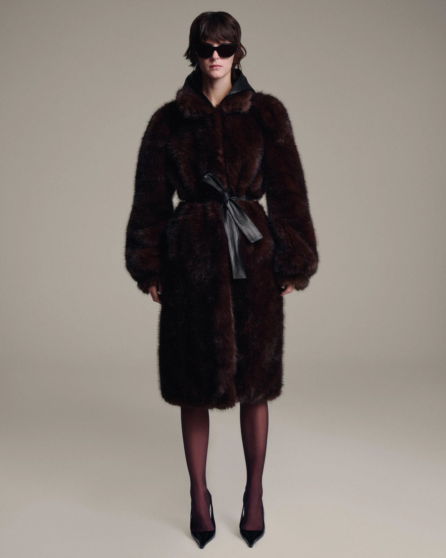A model wearing a dark brown fur coat with a leather hood, a matching waist tie, and dark brown tights with pointed-toe heels, photographed against a solid light beige studio background.