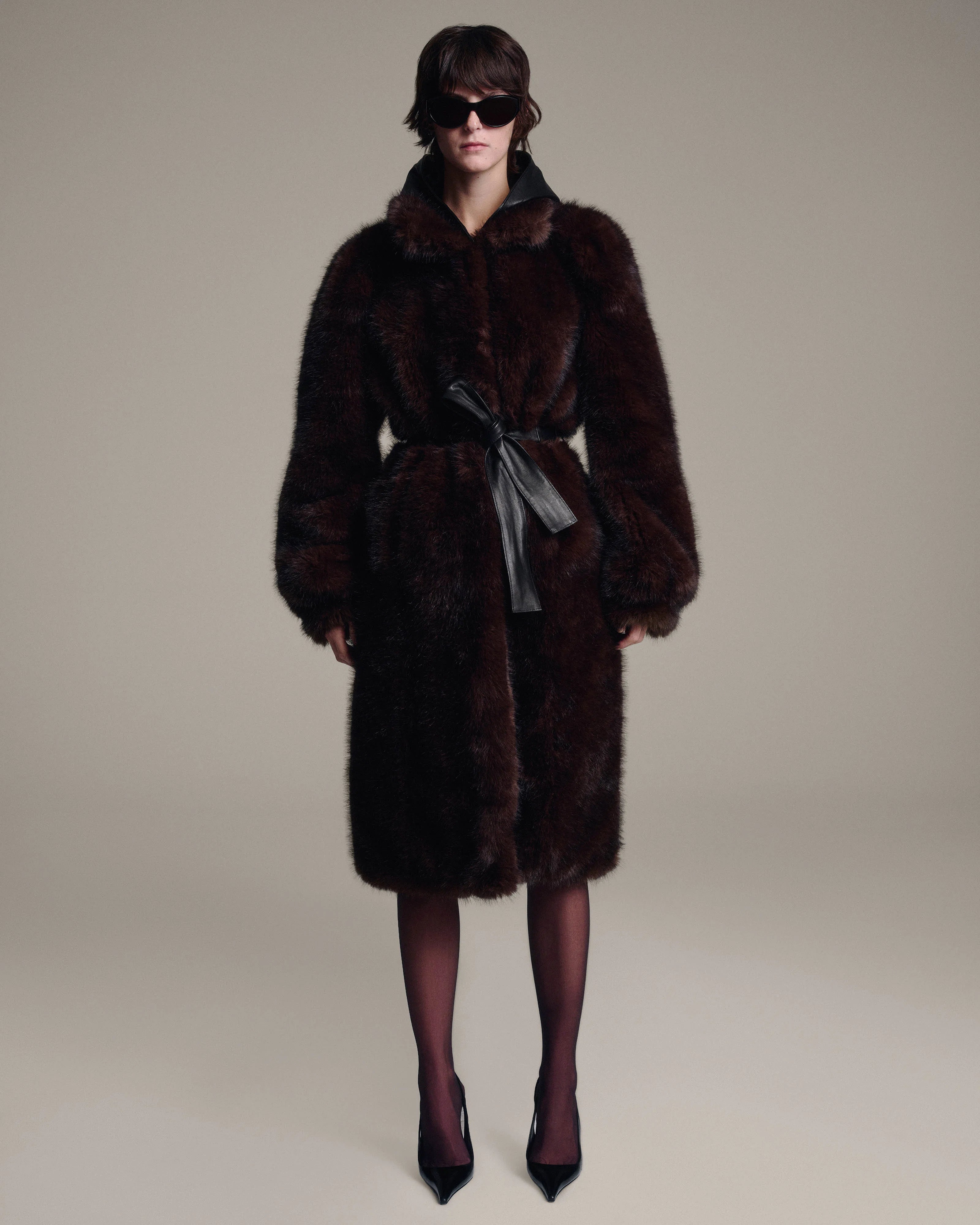 A model wearing a dark brown fur coat with a leather hood, a matching waist tie, and dark brown tights with pointed-toe heels, photographed against a solid light beige studio background.