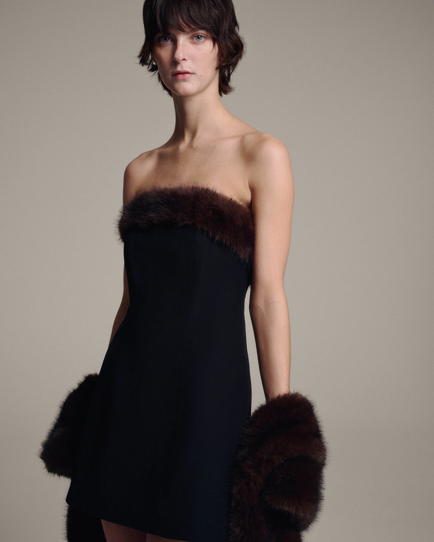 A model wearing a strapless black dress with dark brown fur trim styled with a faux fur shawl, posed against a plain beige studio background.