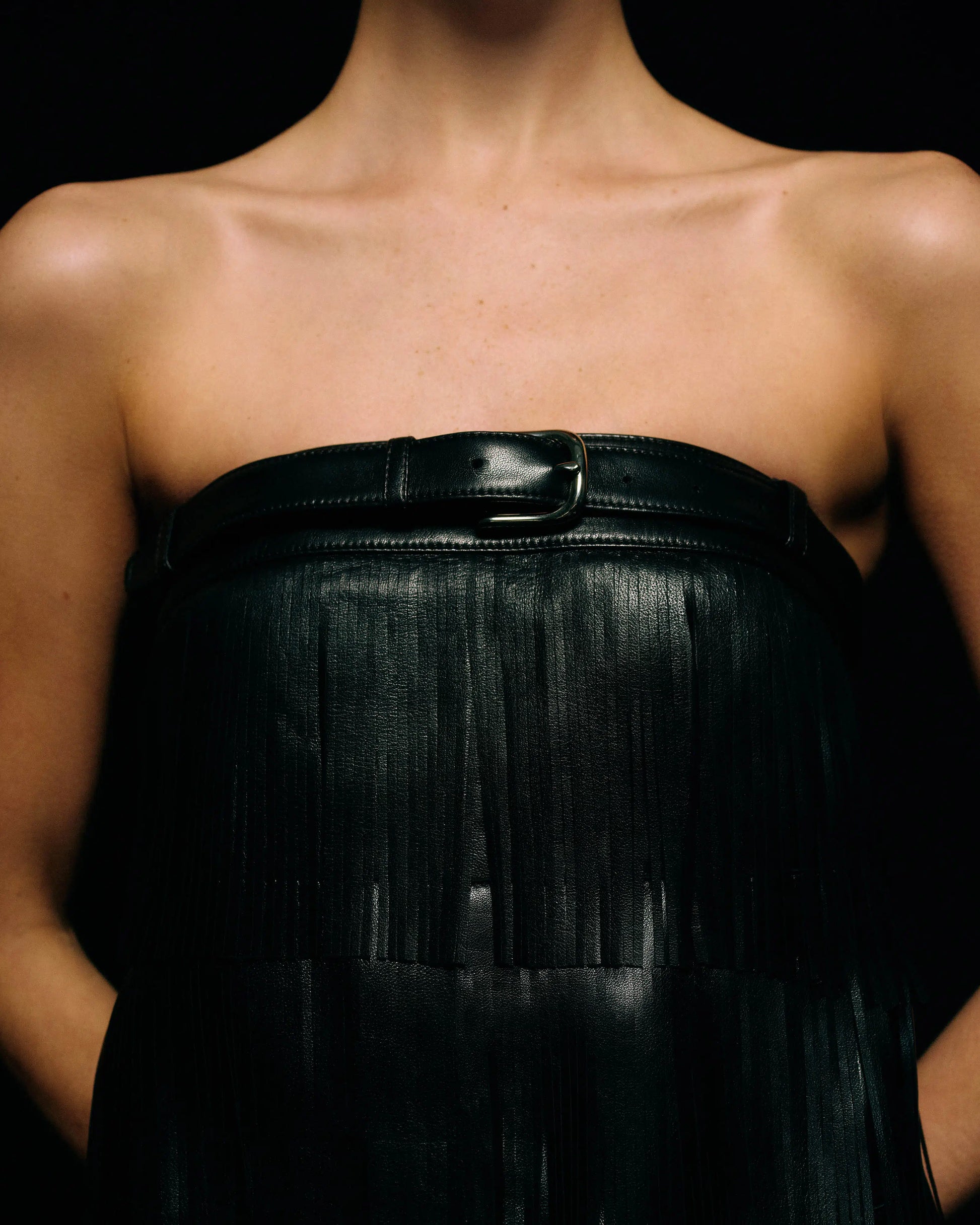 A close-up of a model wearing a black strapless leather fringed dress with a wide black belt across the bust, set against a solid black background.