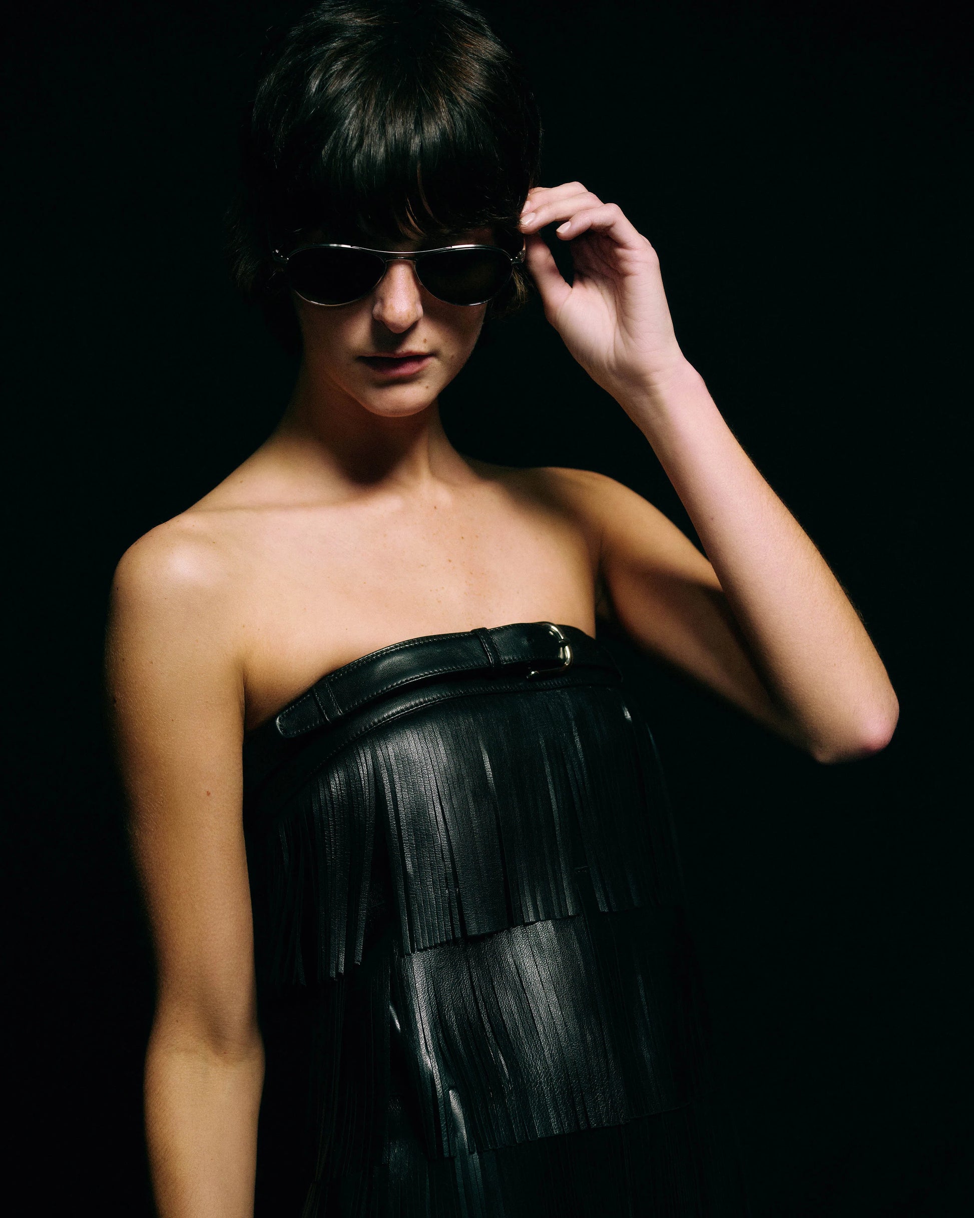 A model wearing a black strapless leather fringe dress and dark sunglasses, posed against a solid black background.