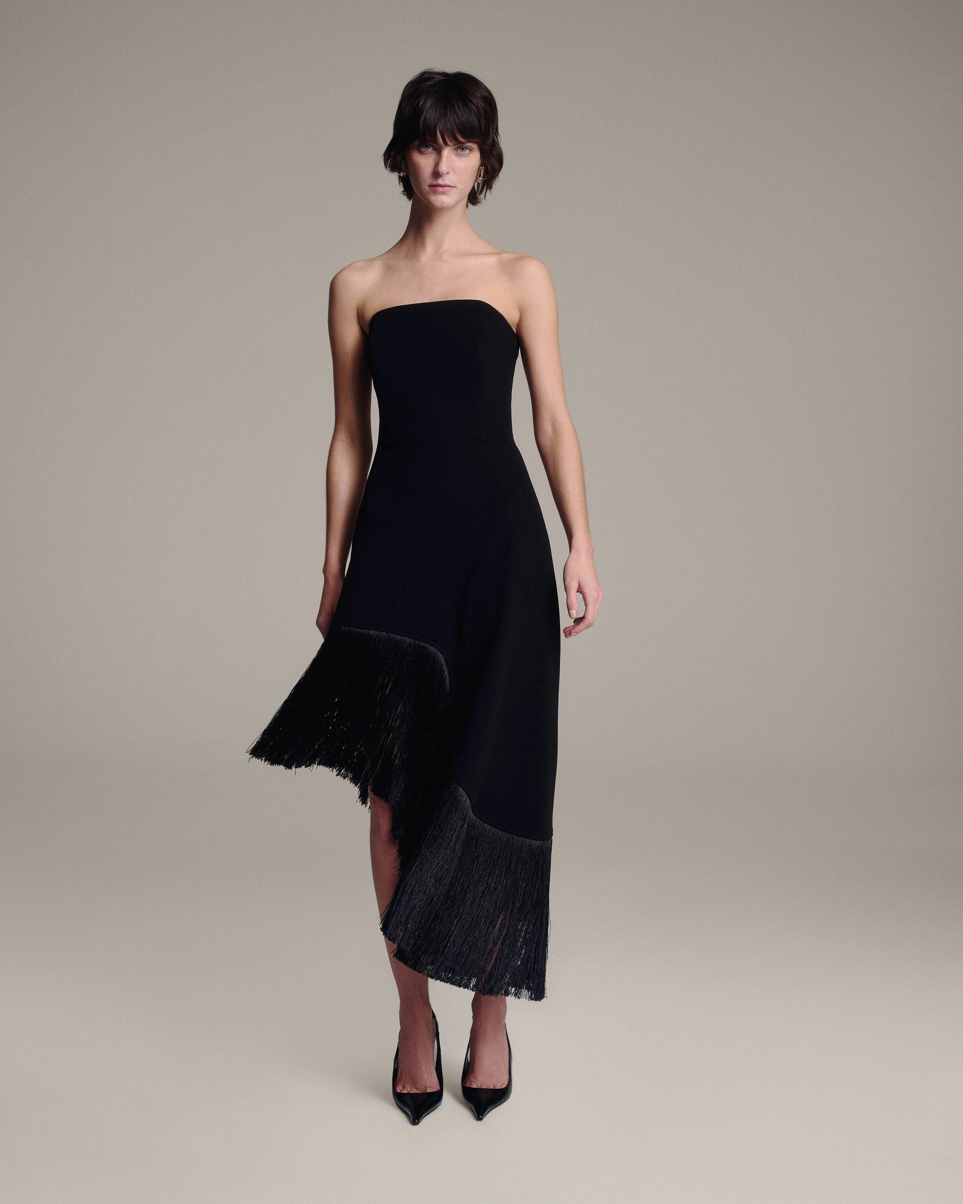 A model wearing a strapless black midi dress with fringe detailing along the hem, styled with black pointed-toe heels against a neutral beige studio background.