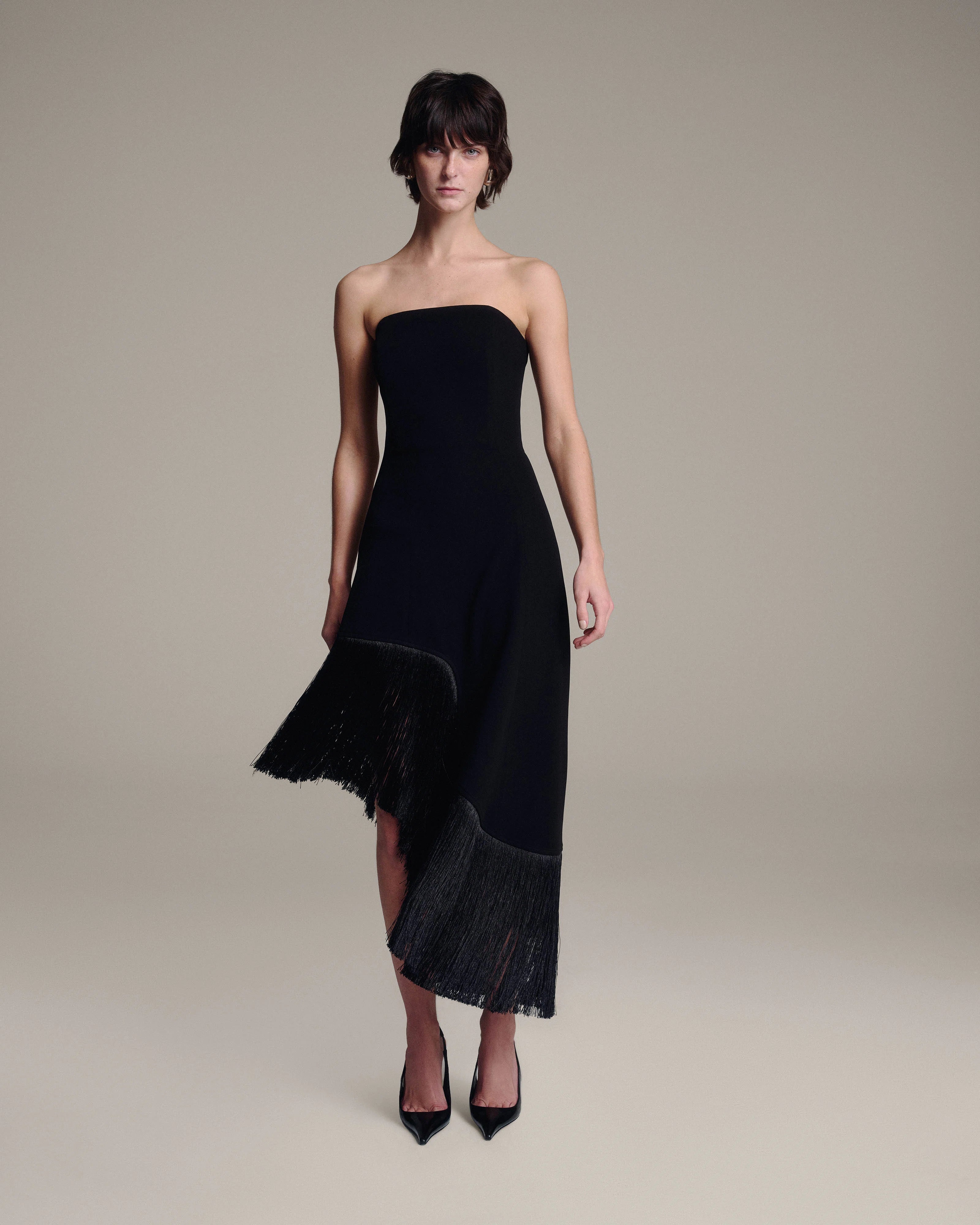 A model wearing a strapless black midi dress with fringe detailing along the hem, styled with black pointed-toe heels against a neutral beige studio background.