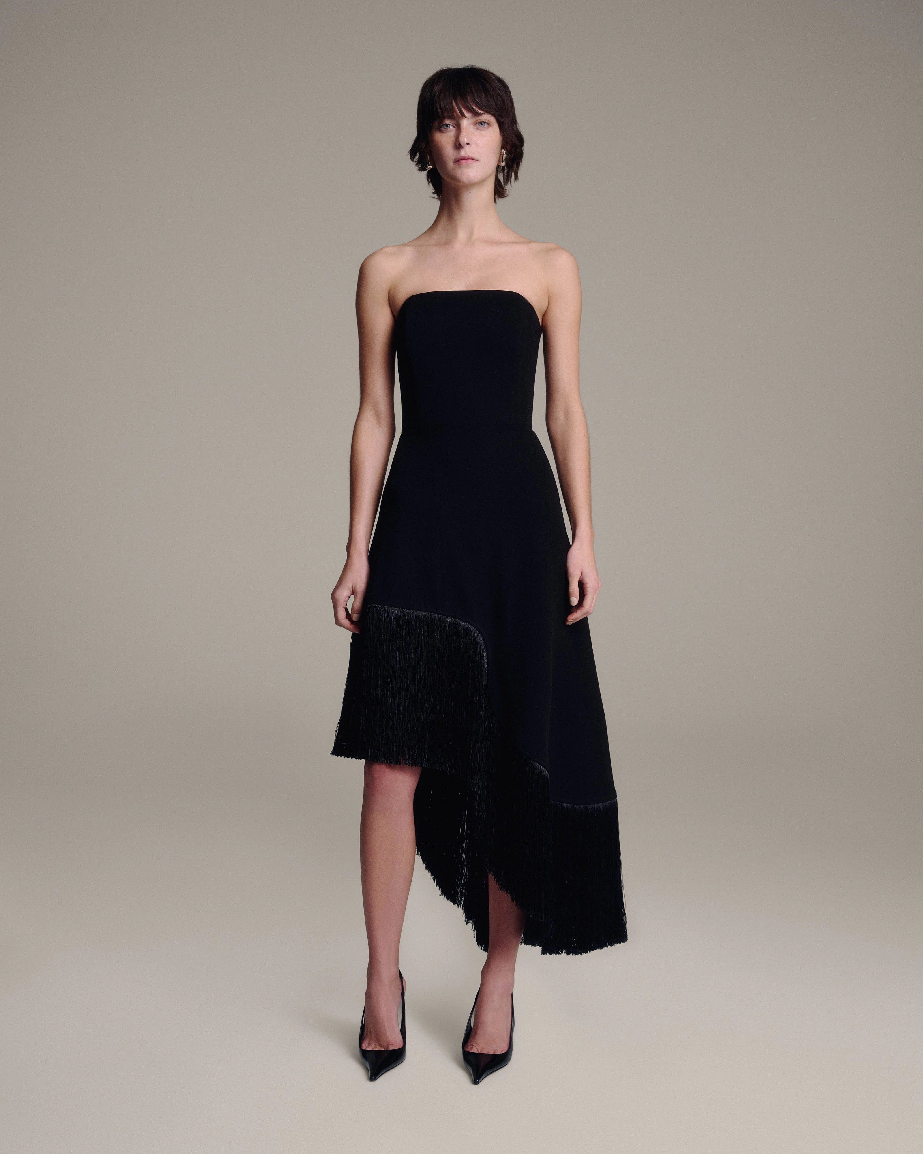 A model wearing a strapless black midi dress with a fringed hem, styled with black pointed-toe heels against a neutral beige studio background.