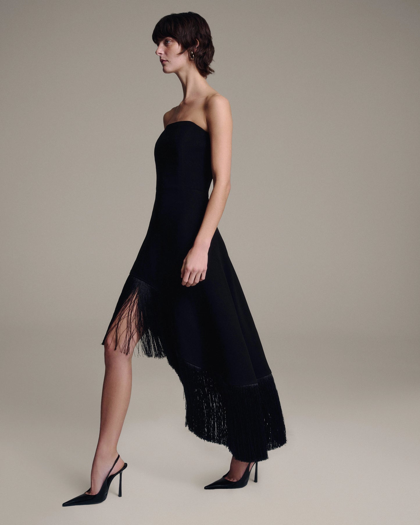 A model wearing a black strapless midi dress with a fringe hem, paired with black pointed-toe heels, set against a solid warm beige studio background.