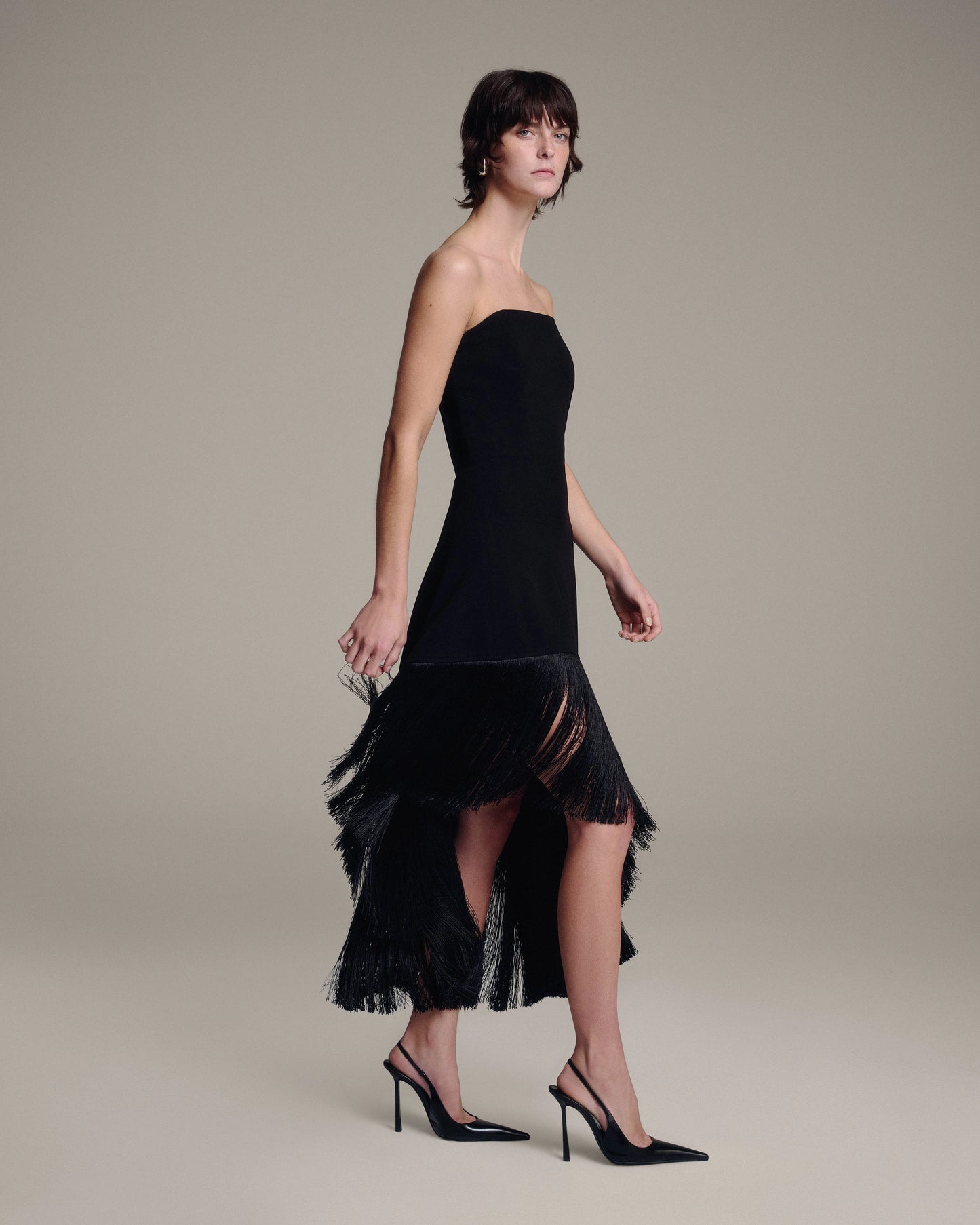 A model wearing a black strapless dress with an asymmetrical, fringed hem and black pointed-toe slingback heels, posed against a solid light gray studio background.