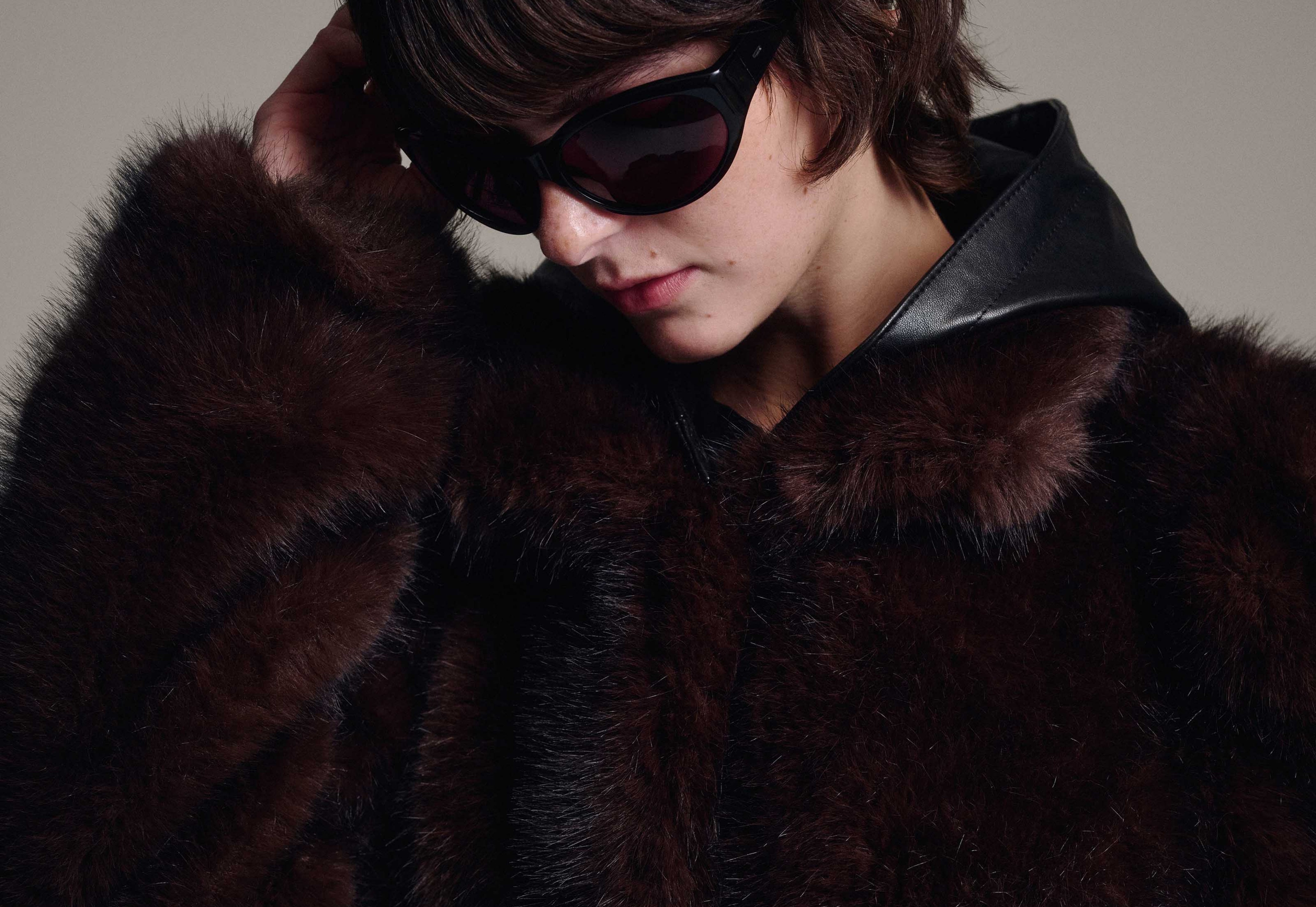A close-up of a model wearing a luxurious dark brown fur coat with a black leather collar, black sunglasses, and a dark brown wig, set against a plain beige background.