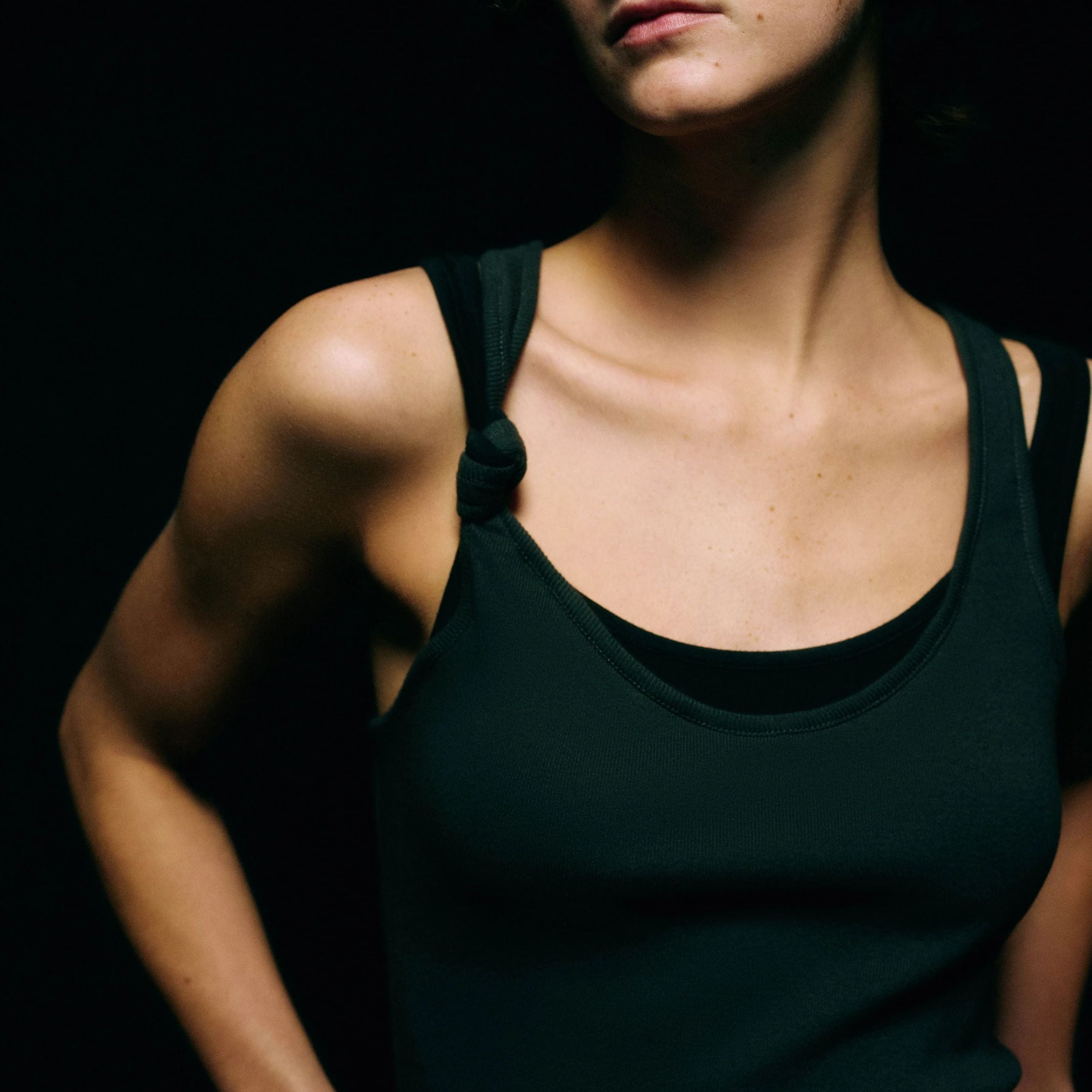 Close-up of the knotted double-strap shoulder detail on a black ribbed tank top against a black background
