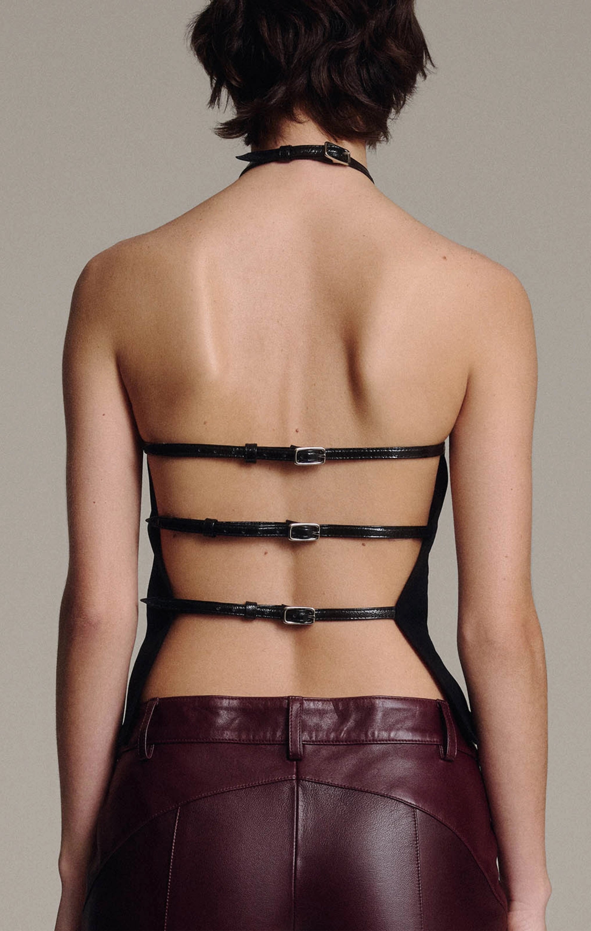 Close-up of the open back of a black halter top with three horizontal buckled leather straps, above the waistband of burgundy leather trousers, against a taupe background